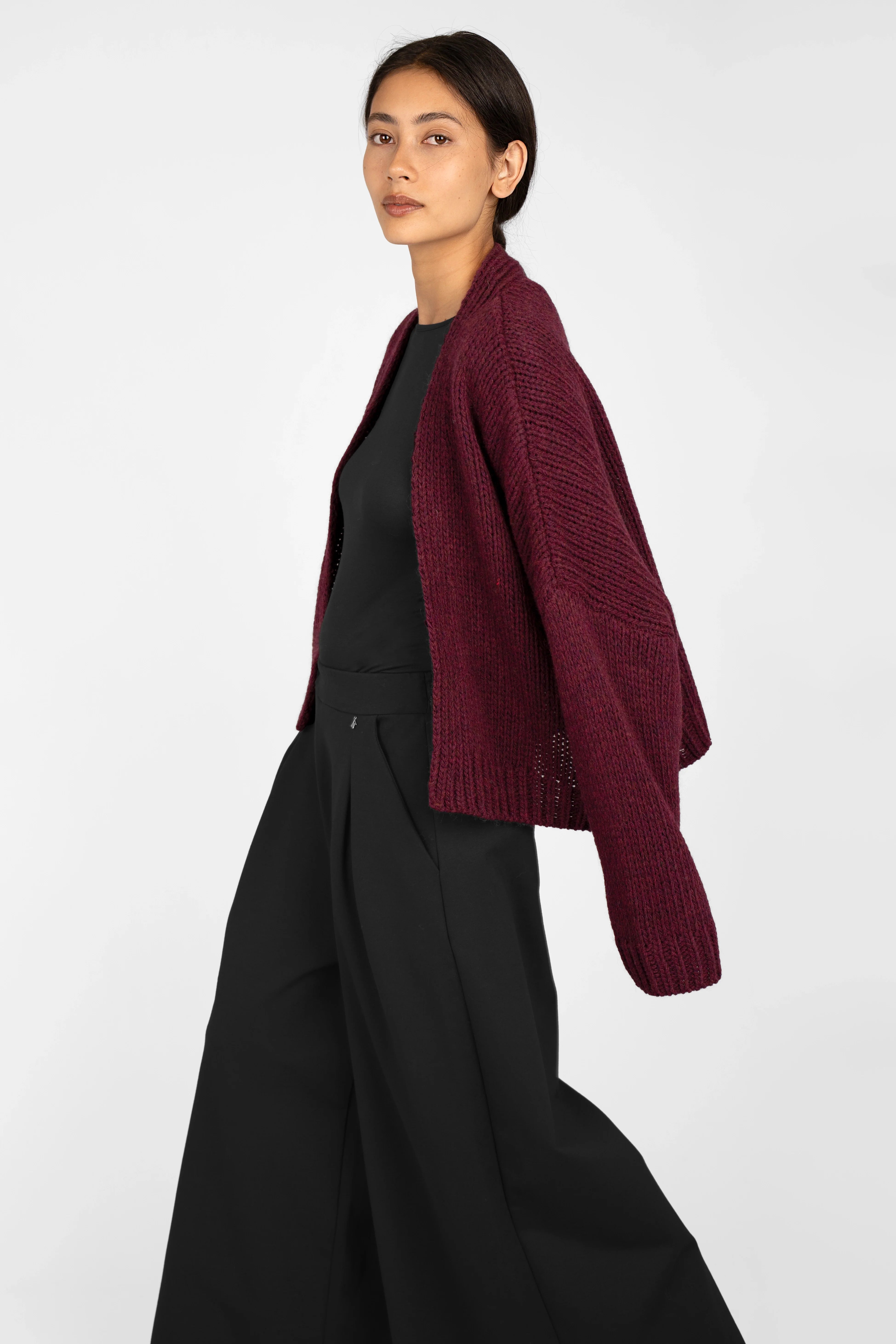 Woman wearing a burgundy ALIXA Cropped Strickcardigan with a shawl collar, loose fit, and ribbed cuffs over a black top and wide-leg black pants.