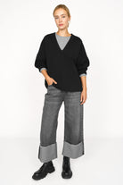 Damen trägt schwarze BERLEY Relaxed V-Neck Bluse aus weichem, formstabilem Jersey mit V-Ausschnitt und überschnittenen Schultern, kombiniert mit grauer Hose und schwarzen Boots