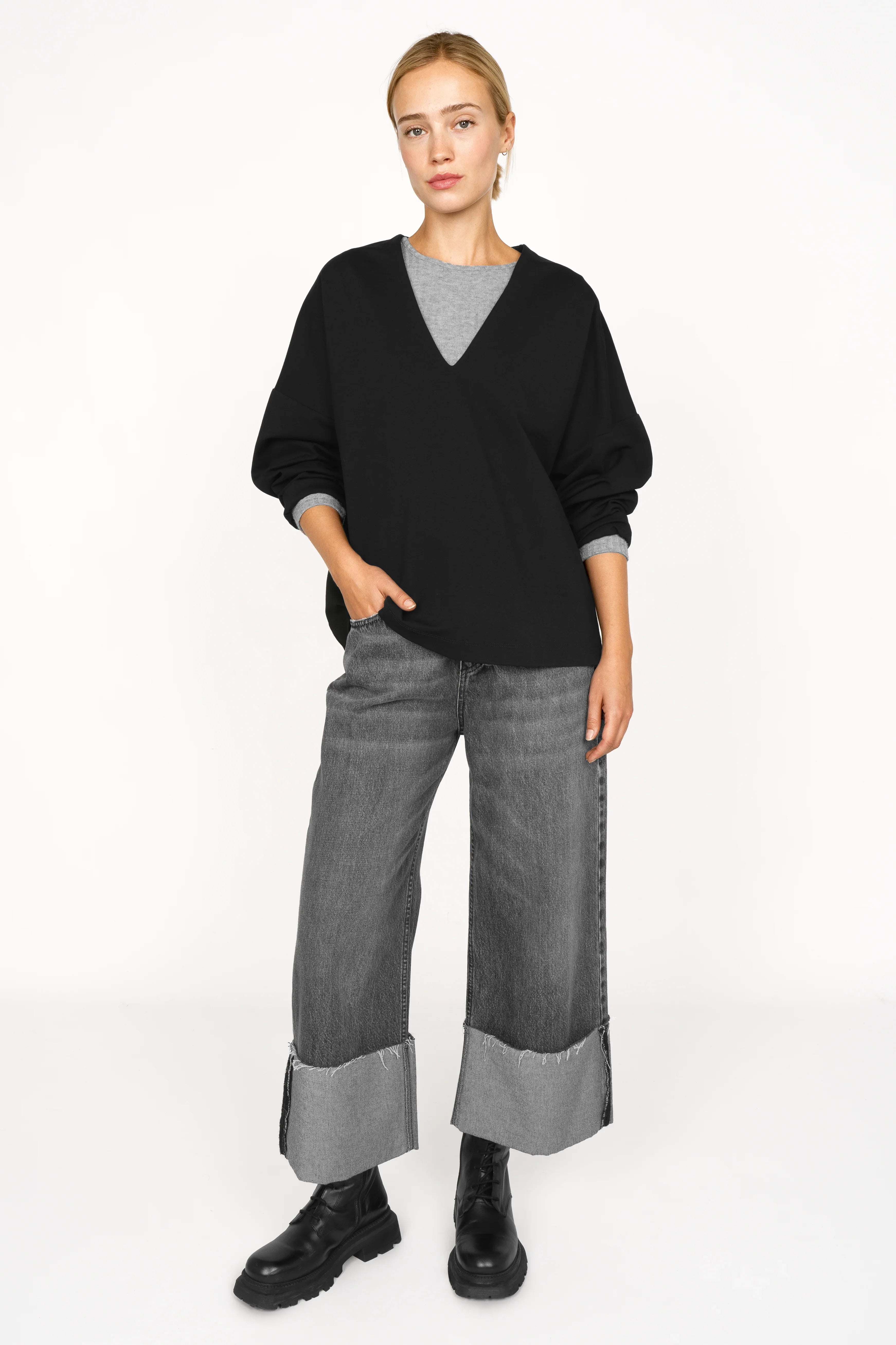 Damen trägt schwarze BERLEY Relaxed V-Neck Bluse aus weichem, formstabilem Jersey mit V-Ausschnitt und überschnittenen Schultern, kombiniert mit grauer Hose und schwarzen Boots