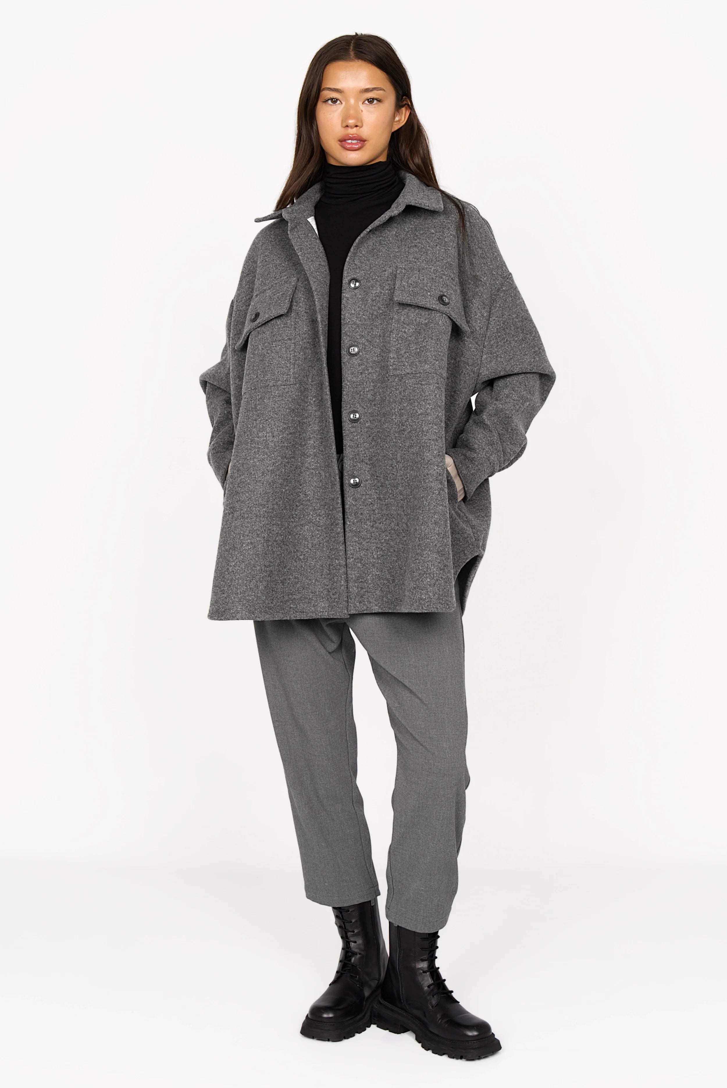 Woman wearing grey BJORA Overshirt with chest pockets and side pockets over black turtleneck and grey pants