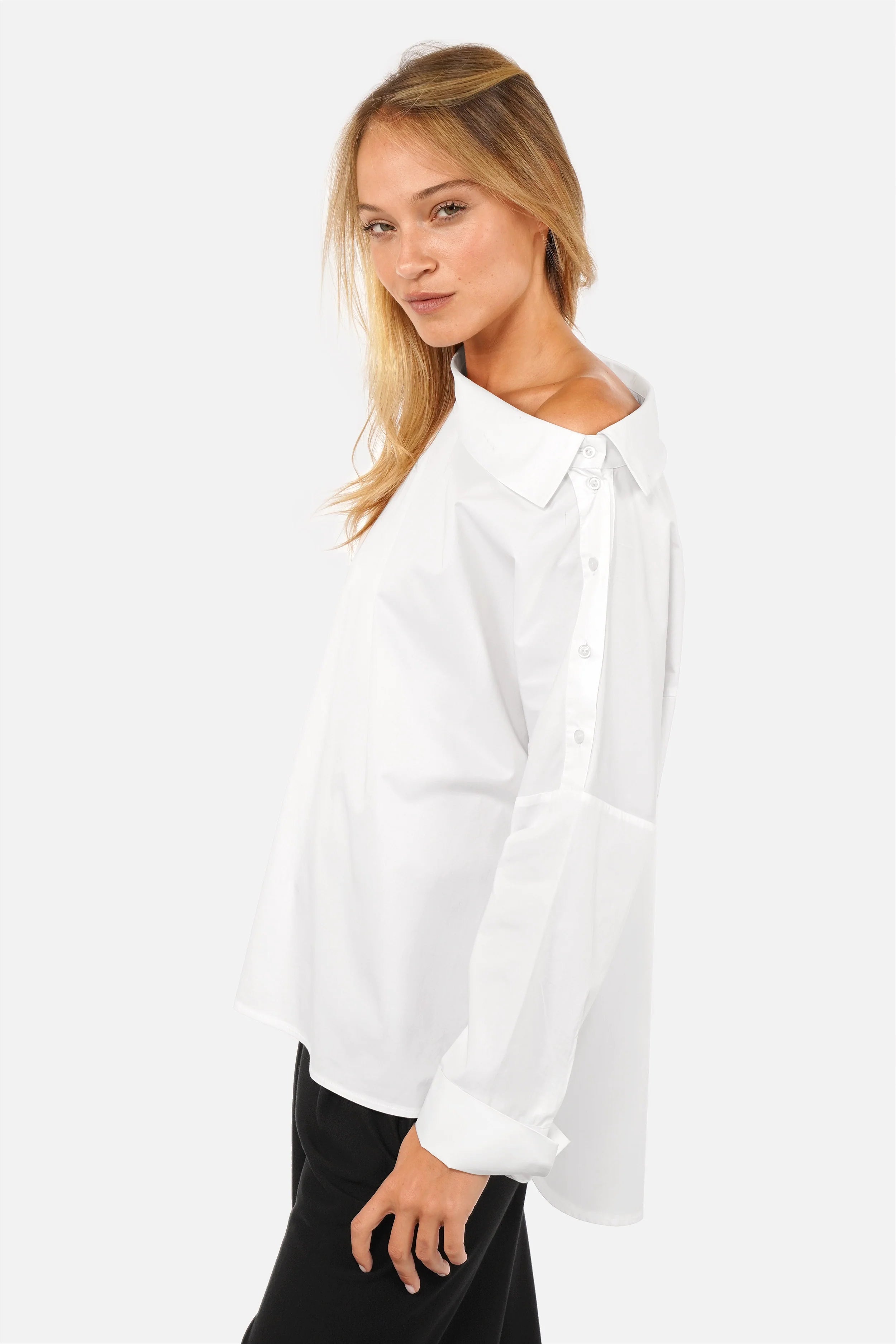 Weißes Modell trägt asymmetrische One Shoulder Bluse aus glattem Baumwollmix mit Knopfleiste und breiten Manschetten