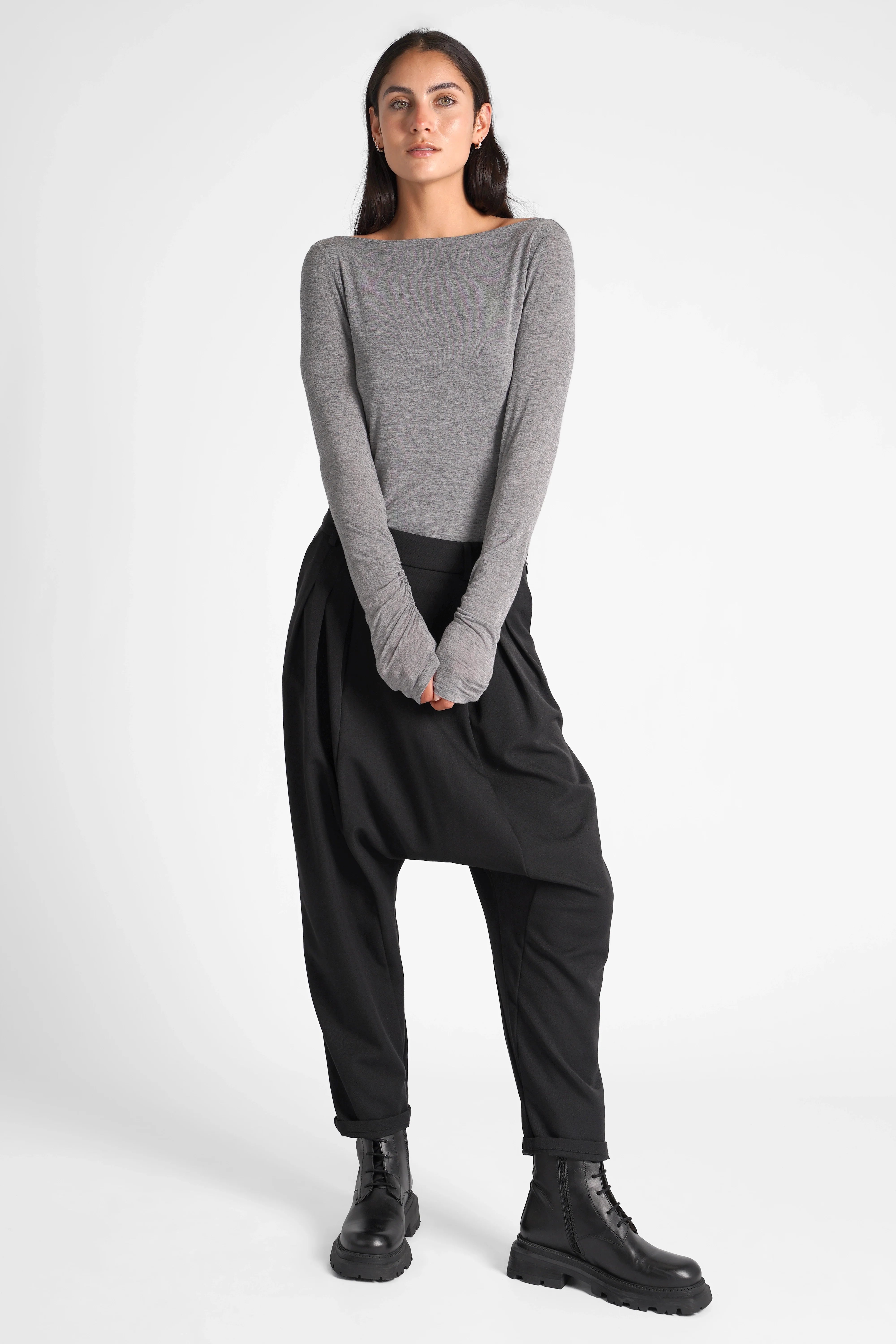 Frau trägt schwarze DEEP Avantgarde-Hose mit tiefem Schritt, seitlicher Knopfleiste und relaxed Fit, kombiniert mit grauem Langarmshirt und schwarzen Stiefeln.