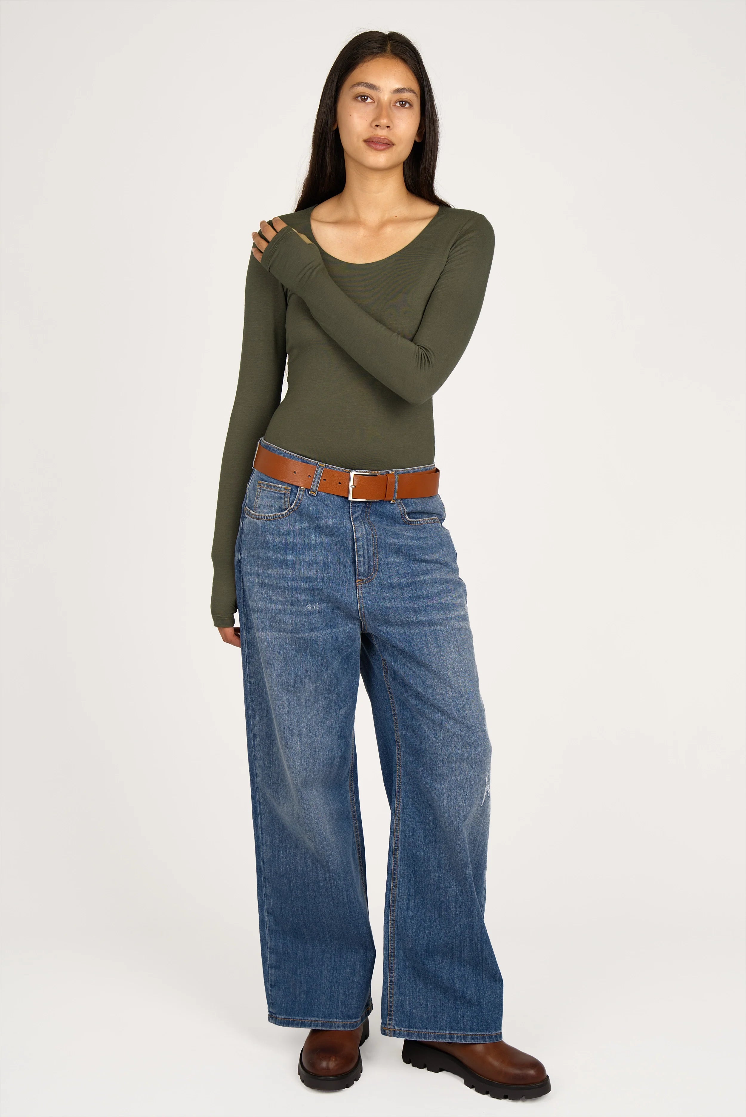 Frau trägt olivgrünes VALUM Longsleeve mit U-Boot-Ausschnitt, extra langen Ärmeln und Daumenlöchern, kombiniert mit weiter blauer Jeans und braunen Schuhen.