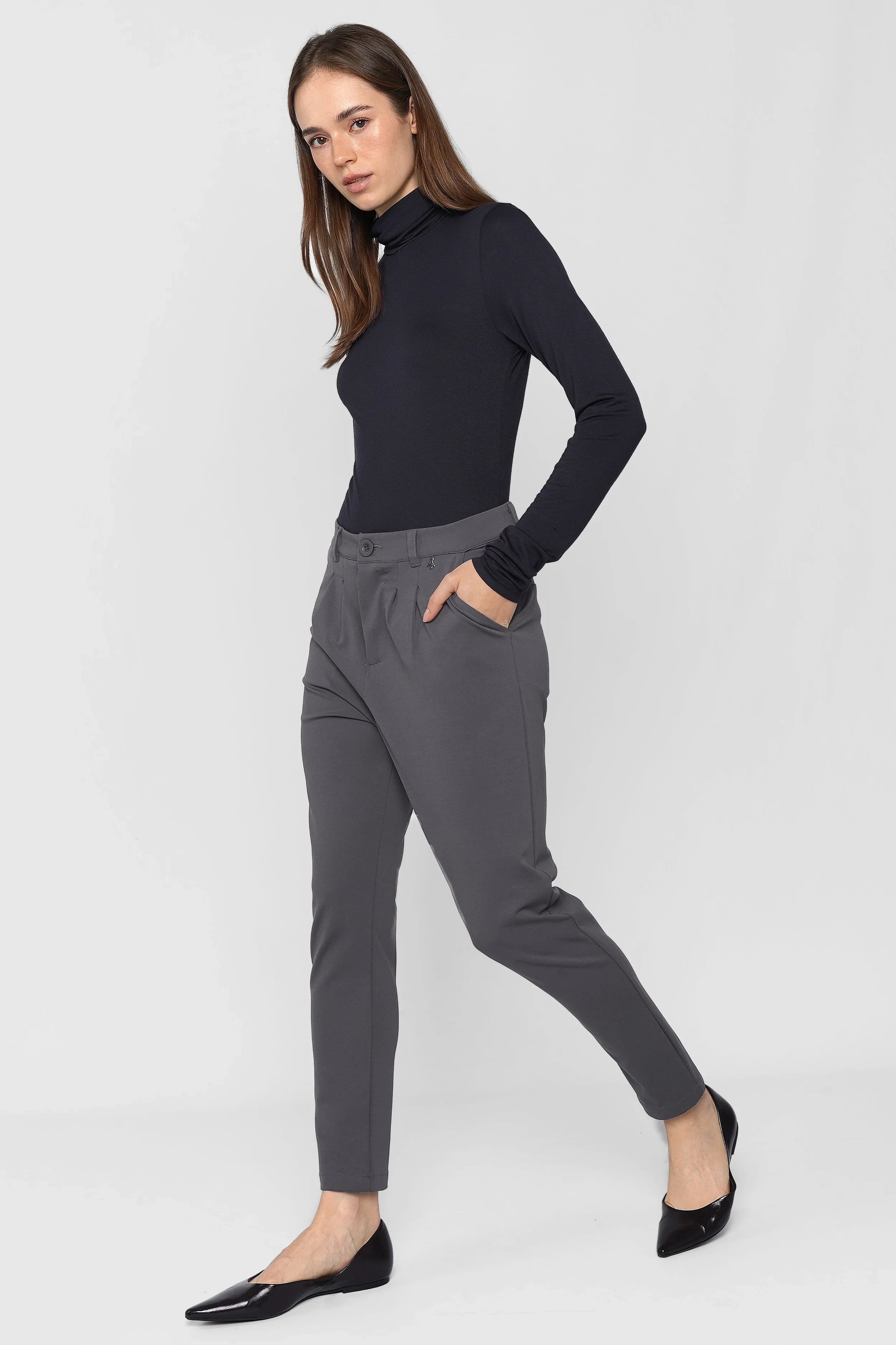 Woman wearing HEELENA tapered pants in gray with black long-sleeve top and flats standing on gray background