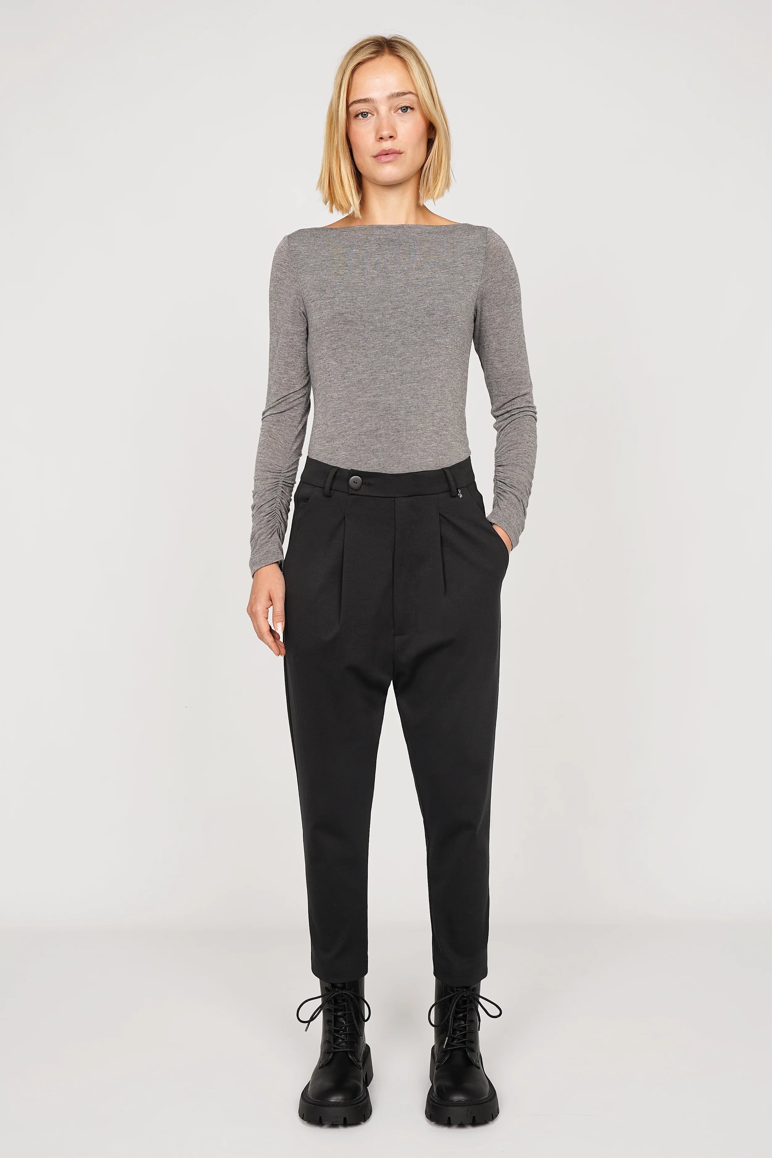 Frau trägt schwarze JALDI Cropped Tapered Hose mit schmal zulaufendem Bein und elastischem Bund, kombiniert mit grauem Langarmshirt und schwarzen Stiefeln.