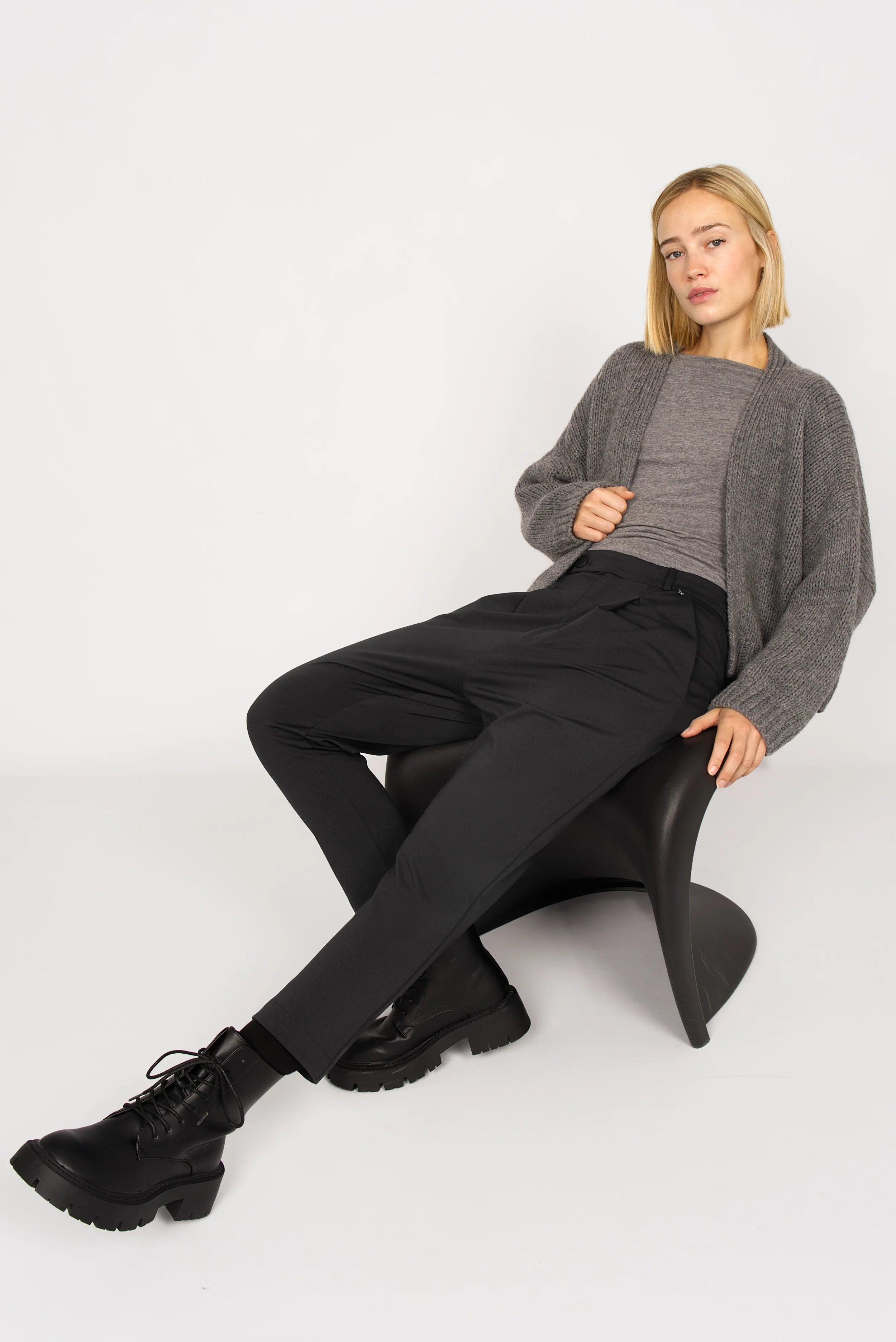 Frau trägt schwarze JALDI Cropped Tapered Hose mit engem Beinverlauf und elastischem Bund, kombiniert mit grauem Pullover und Boots