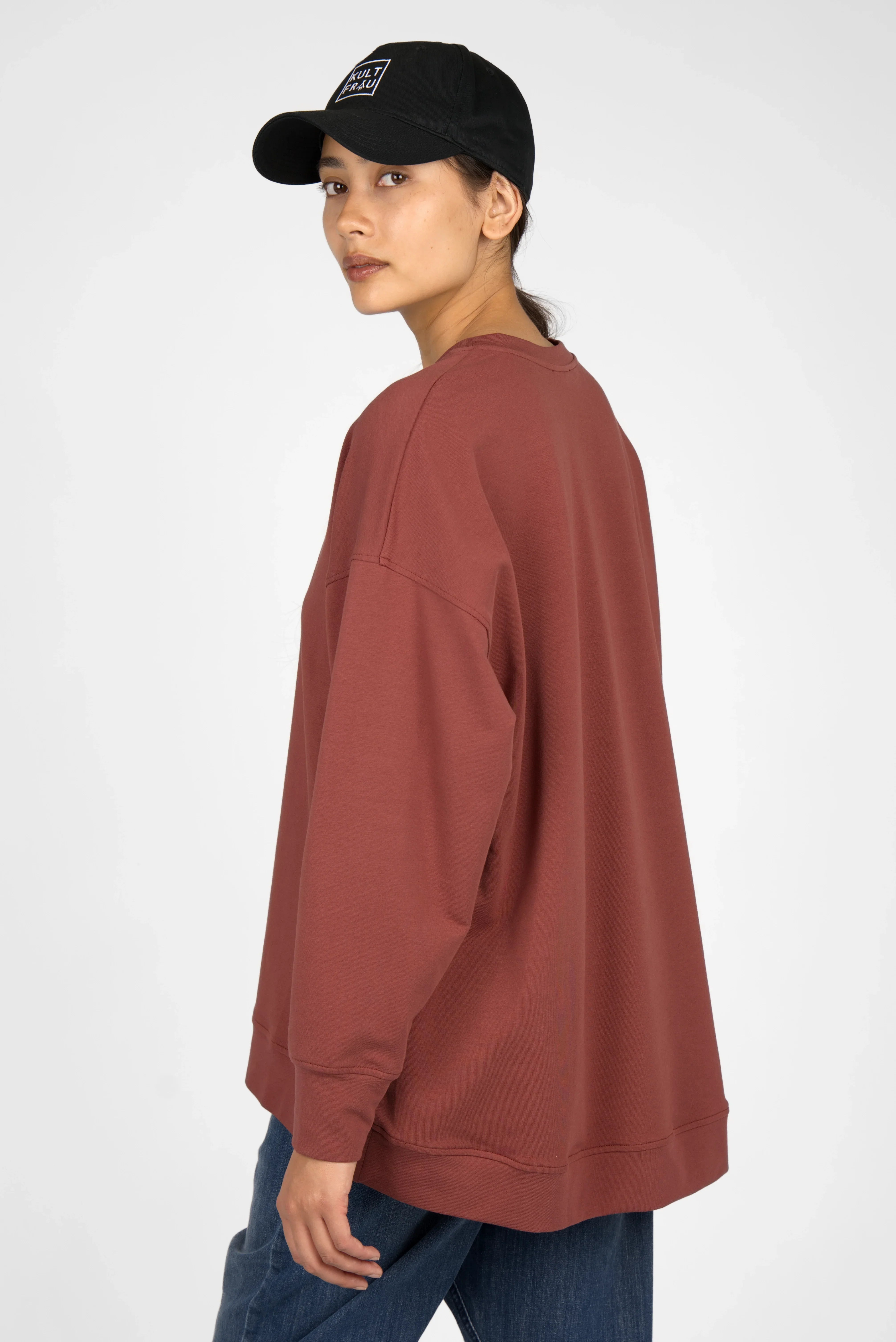 Woman wearing brown EIREL oversize sweater with dropped shoulders and round neckline, paired with a black cap and blue jeans.