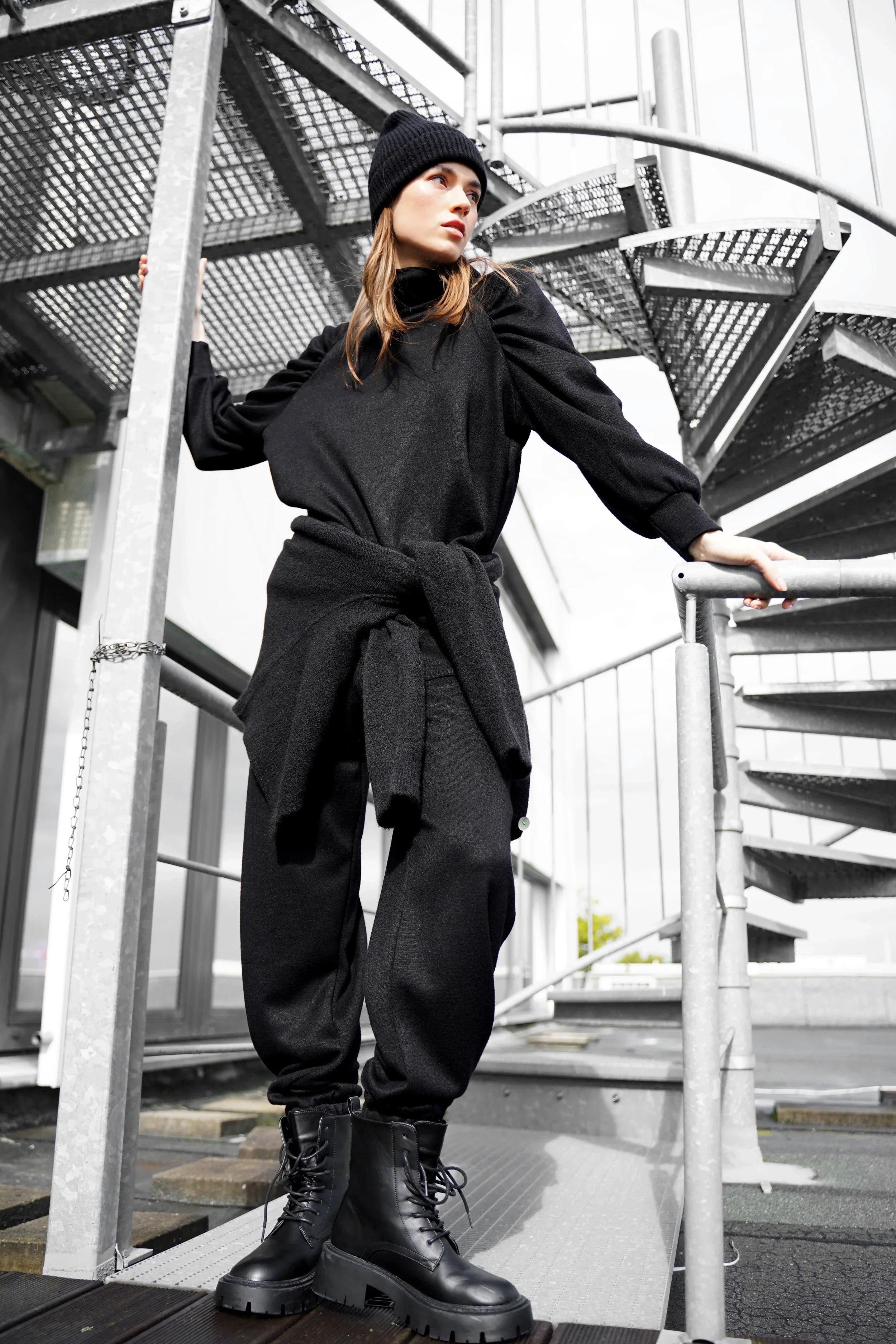 Woman wearing black FRELA Jogpant aus Feinstrick with elastic waistband and ribbed cuffs, styled with black boots and beanie on outdoor staircase