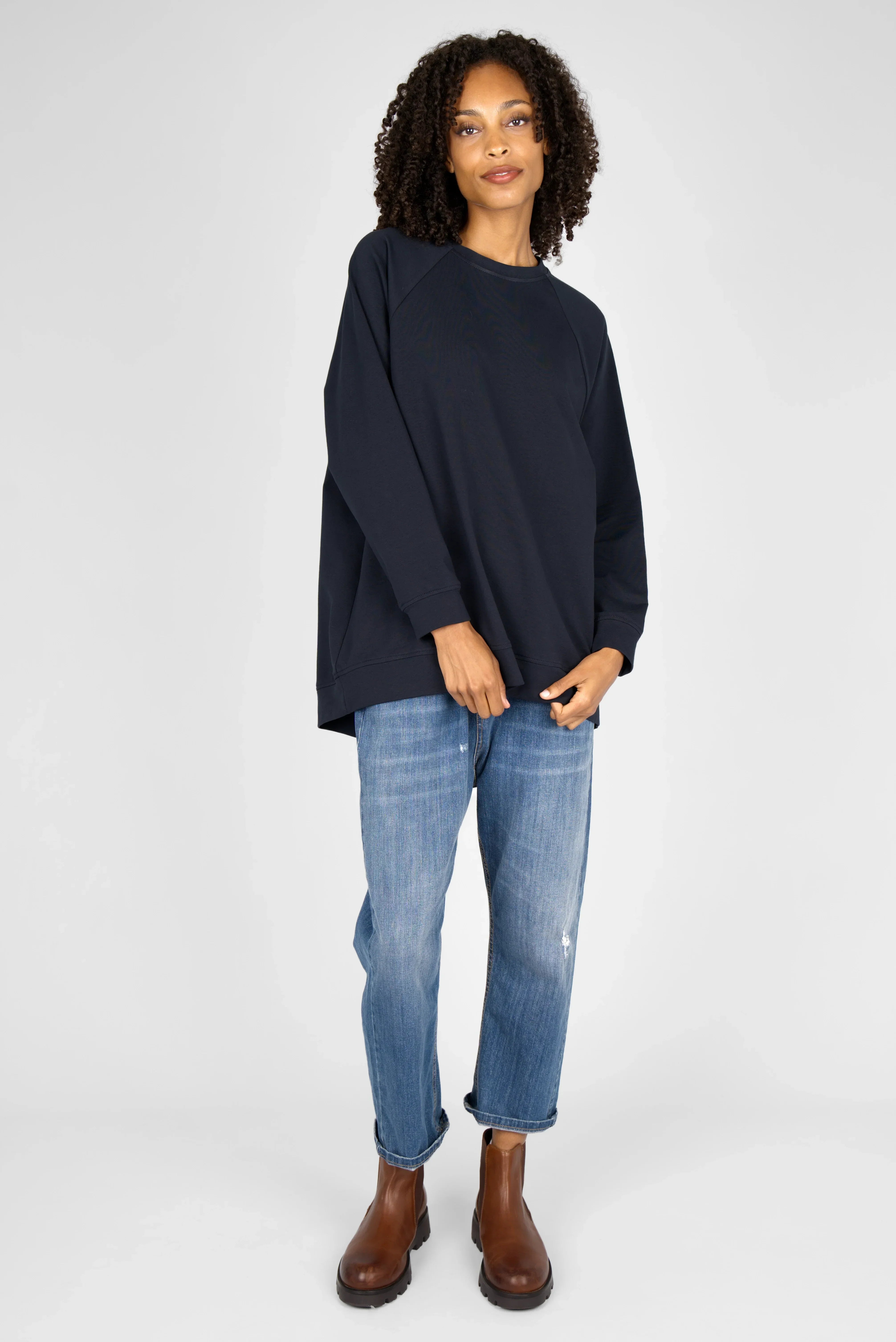 Woman wearing navy GRACEY.K Sweater with raglan sleeves and rounded hem, paired with blue jeans and brown boots.
