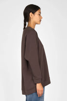 Side view of a woman wearing a brown GRACEY.K Sweater with oversized fit, round neckline, and longer back hem.