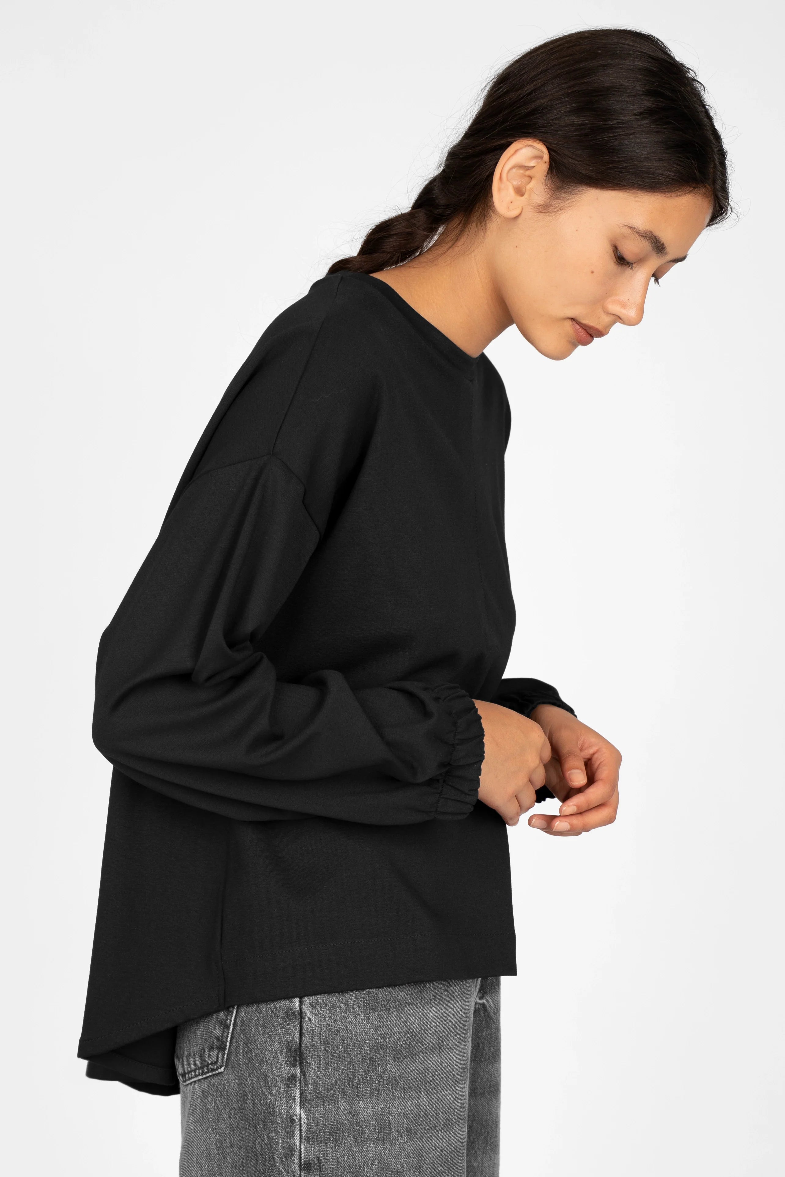 Woman wearing black HAGLA Shirt with round neckline, relaxed fit, elastic cuffs, and longer back hem paired with gray jeans against white background