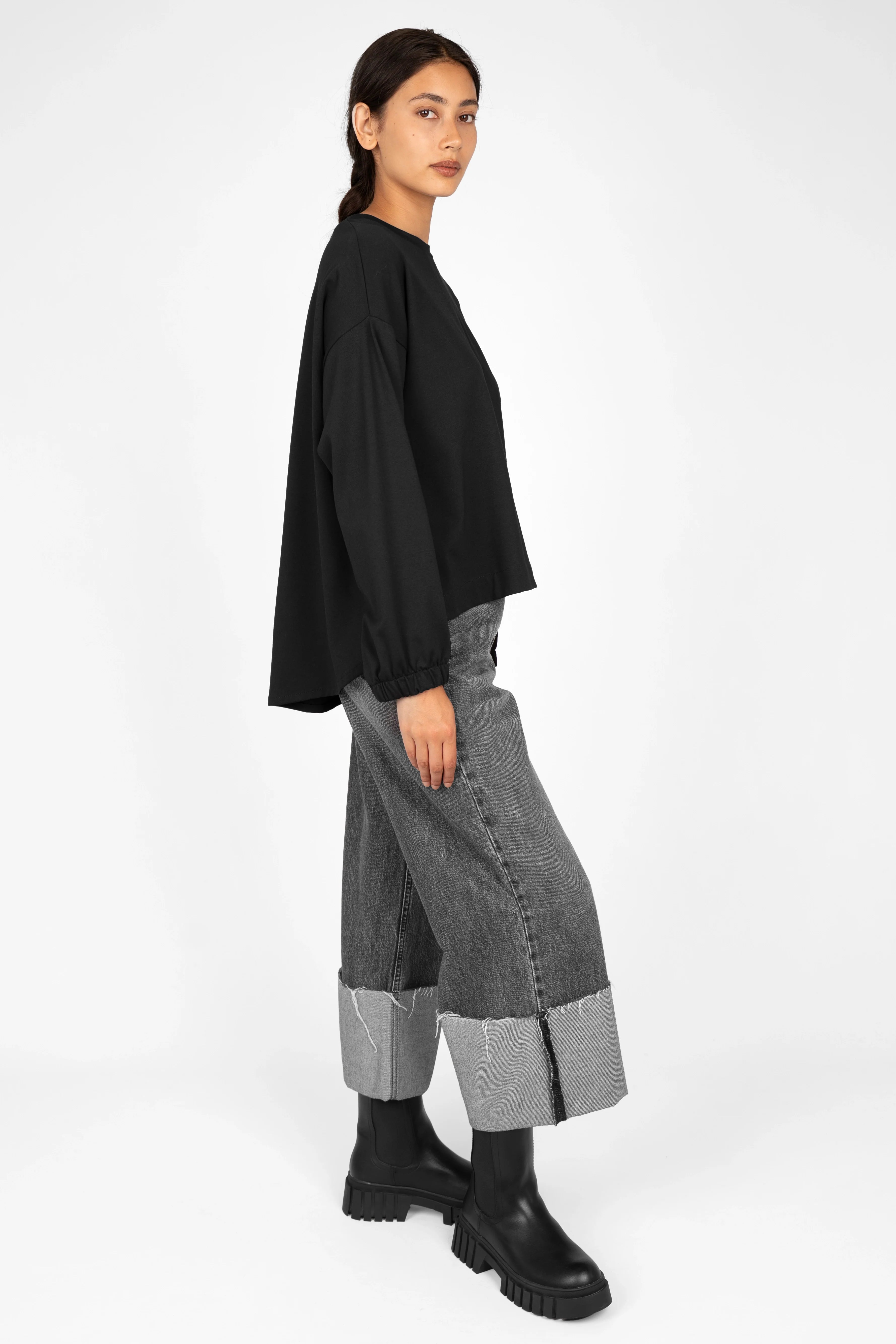 Woman wearing black HAGLA Shirt with relaxed fit, round neckline, elastic cuffs, and longer back hem paired with cuffed gray jeans and black boots, side view.