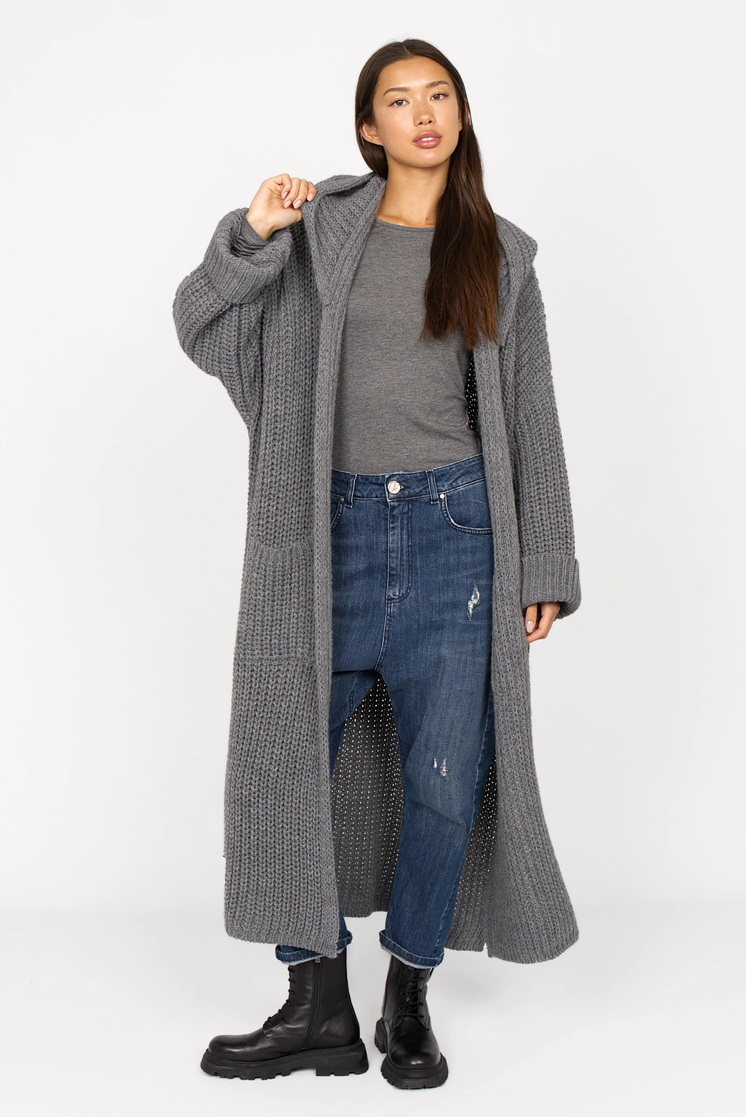 Frau trägt JEANY Baggy Jeans mit lockerem Sitz, mittlerer blauer Waschung und Used-Effekten, kombiniert mit grauem Pullover und langem grauem Strickmantel