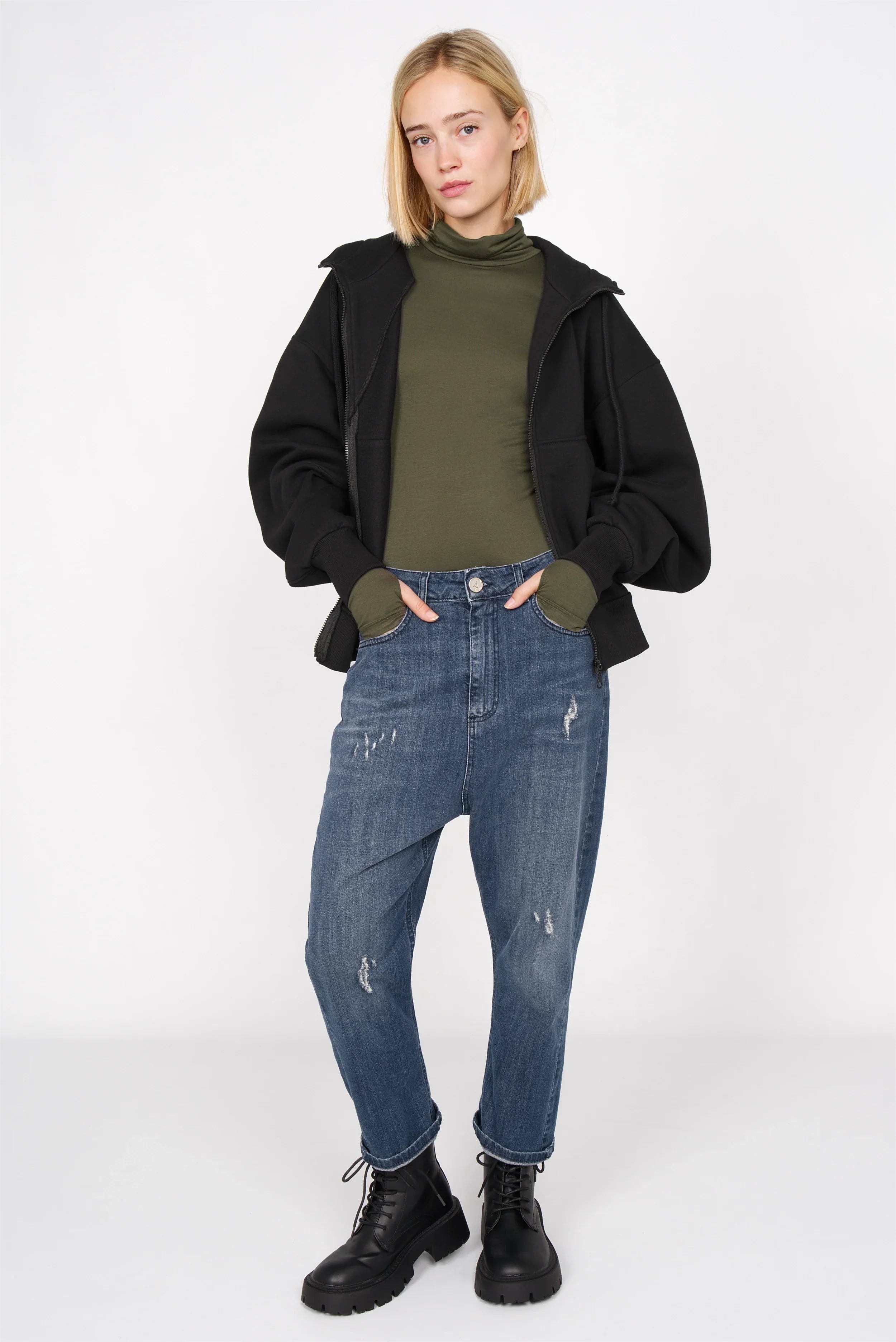 Frau trägt olivgrünes KABEA Longsleeve mit Rollkragen und Daumenlöchern zu Jeans und schwarzen Boots