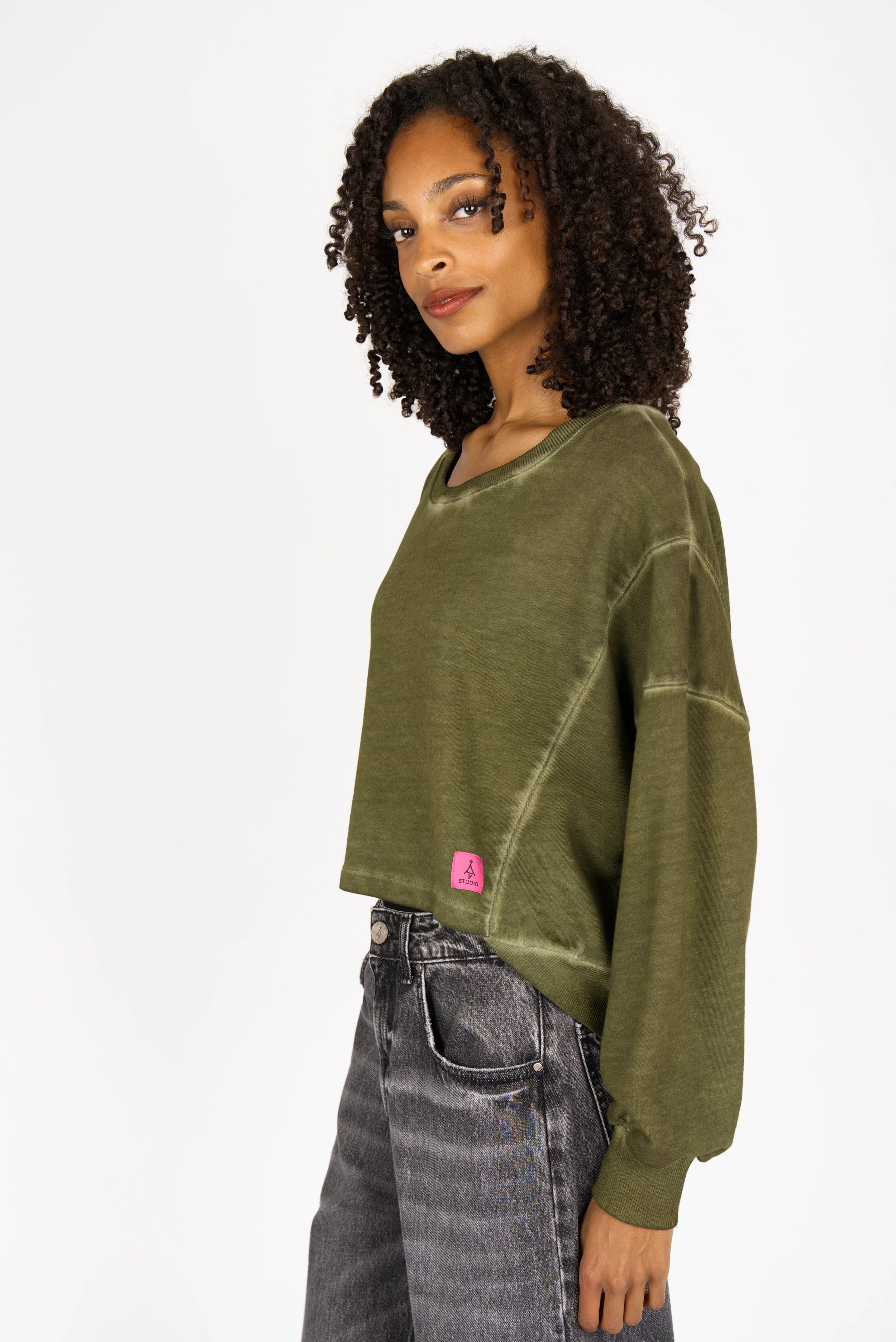 Woman wearing olive green KERY Cropped Sweater with oversized fit, U-boat neckline, and pink STUDIO logo patch.
