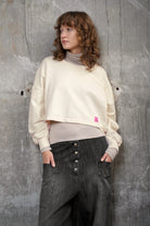 KERY Cropped Sweater in Oversized Passform mit U-Boot-Ausschnitt und pinkem Studio Patch vor grauer Wand