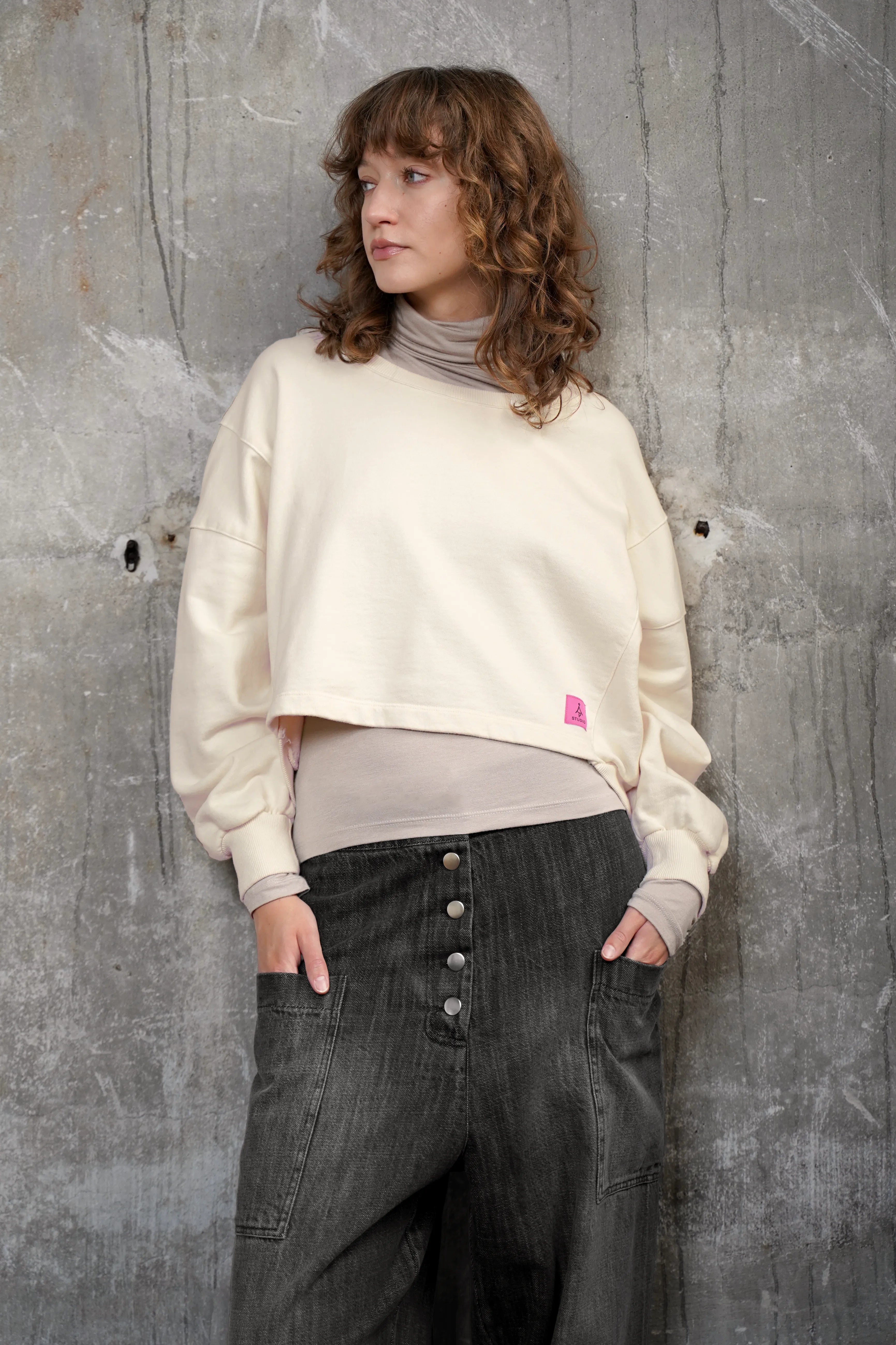KERY Cropped Sweater in Oversized Passform mit U-Boot-Ausschnitt und pinkem Studio Patch vor grauer Wand
