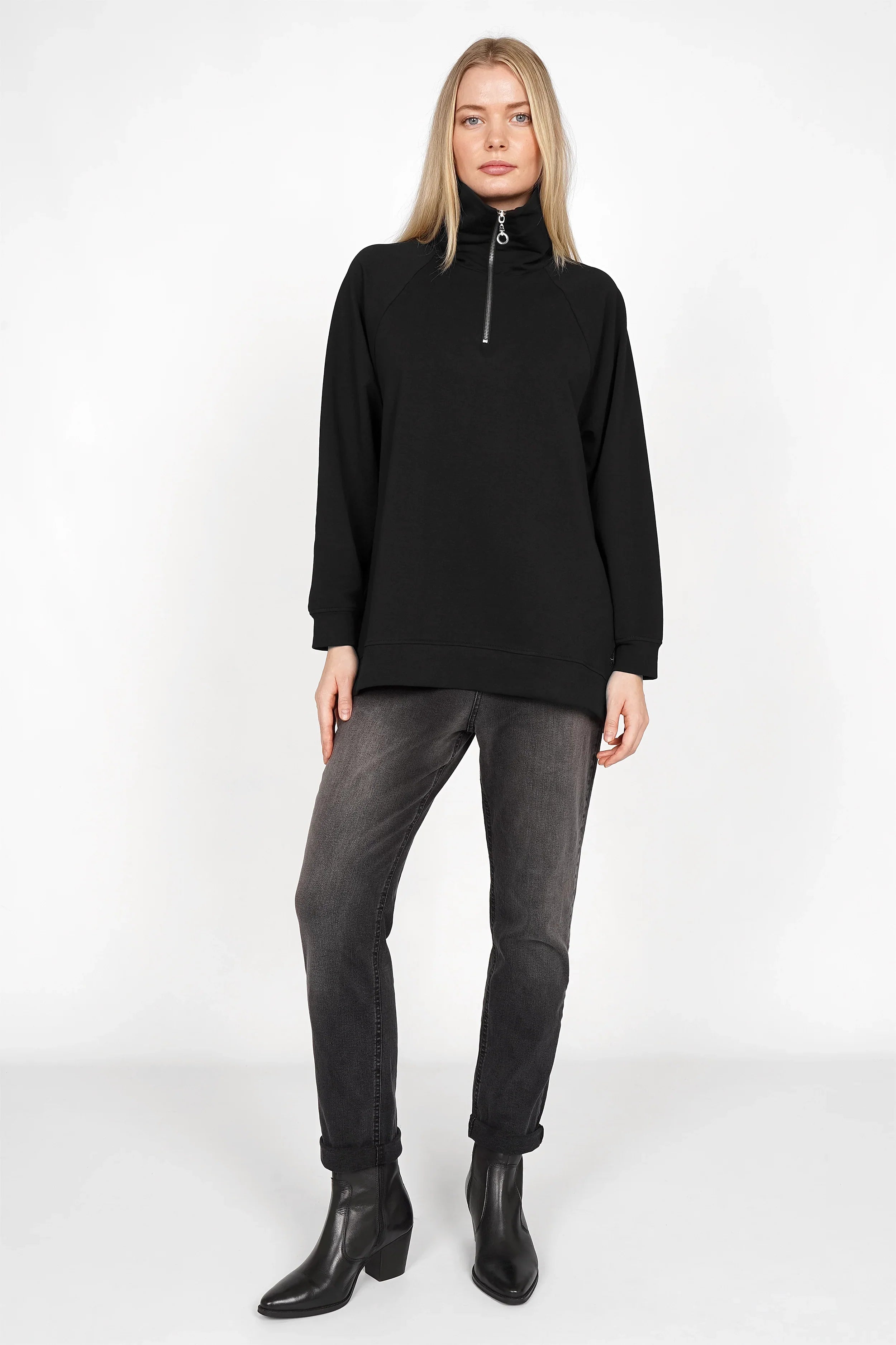 Woman wearing black Lenia Troyer-Oversize Sweater with half-zip and raglan sleeves, paired with dark jeans and black boots.