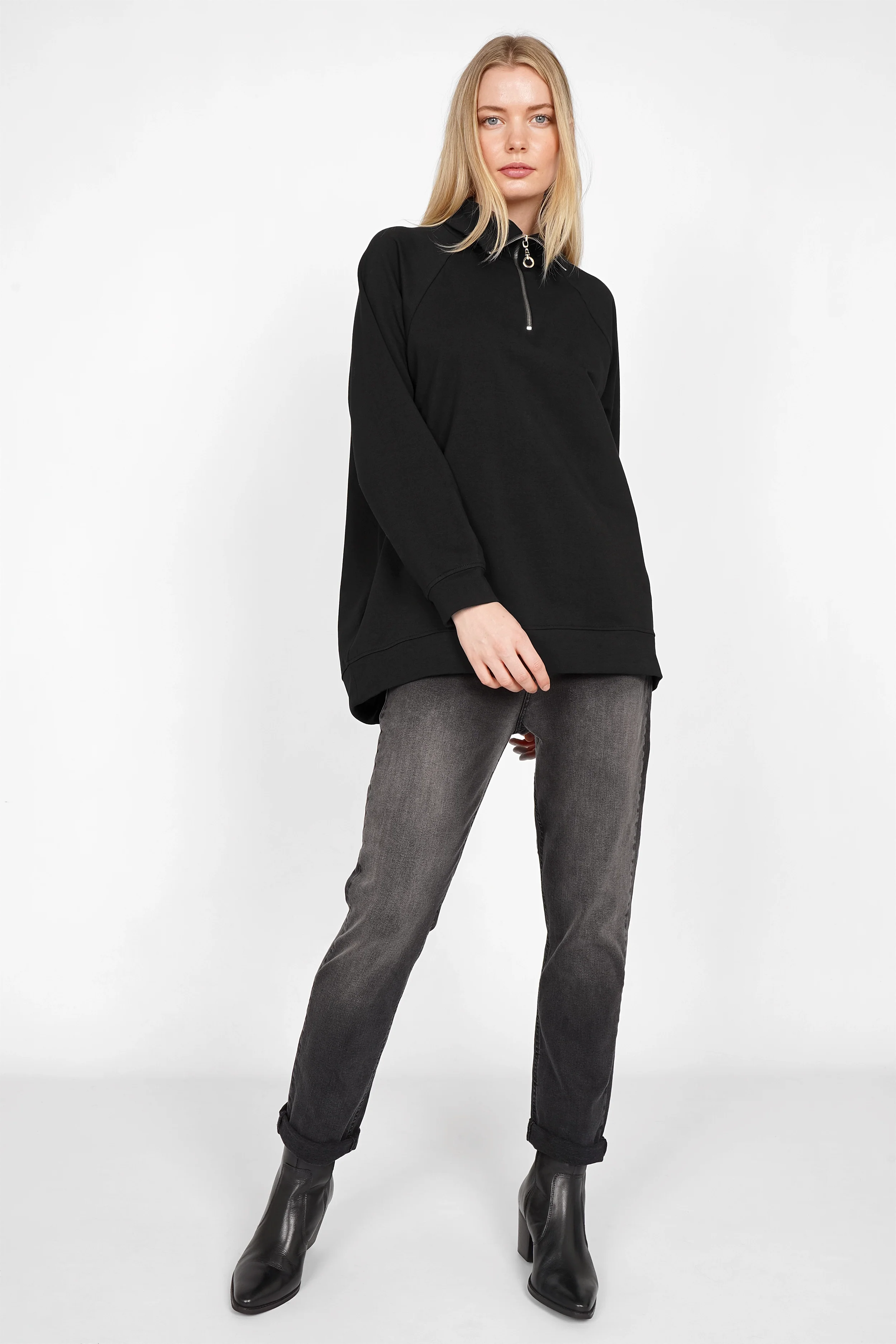 Frau trägt schwarzen LENIA Troyer-Oversize Sweater mit halbem Metallzipper und Raglanärmeln, kombiniert mit grauen Jeans und schwarzen Stiefeletten.
