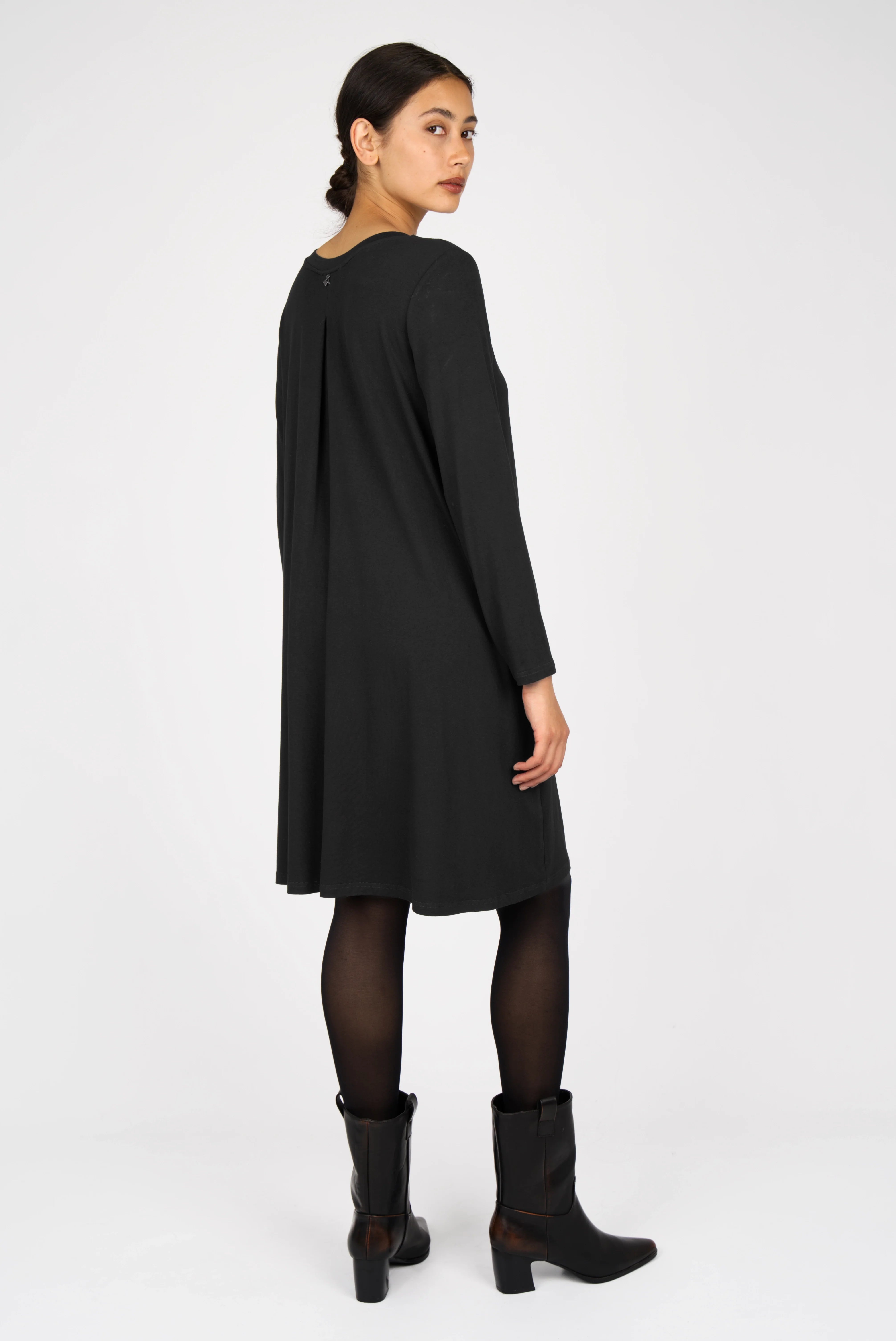 Woman wearing a black LYLLDA A-Linie Kleid dress with long sleeves, a round neckline, and a back pleat, paired with black tights and ankle boots.