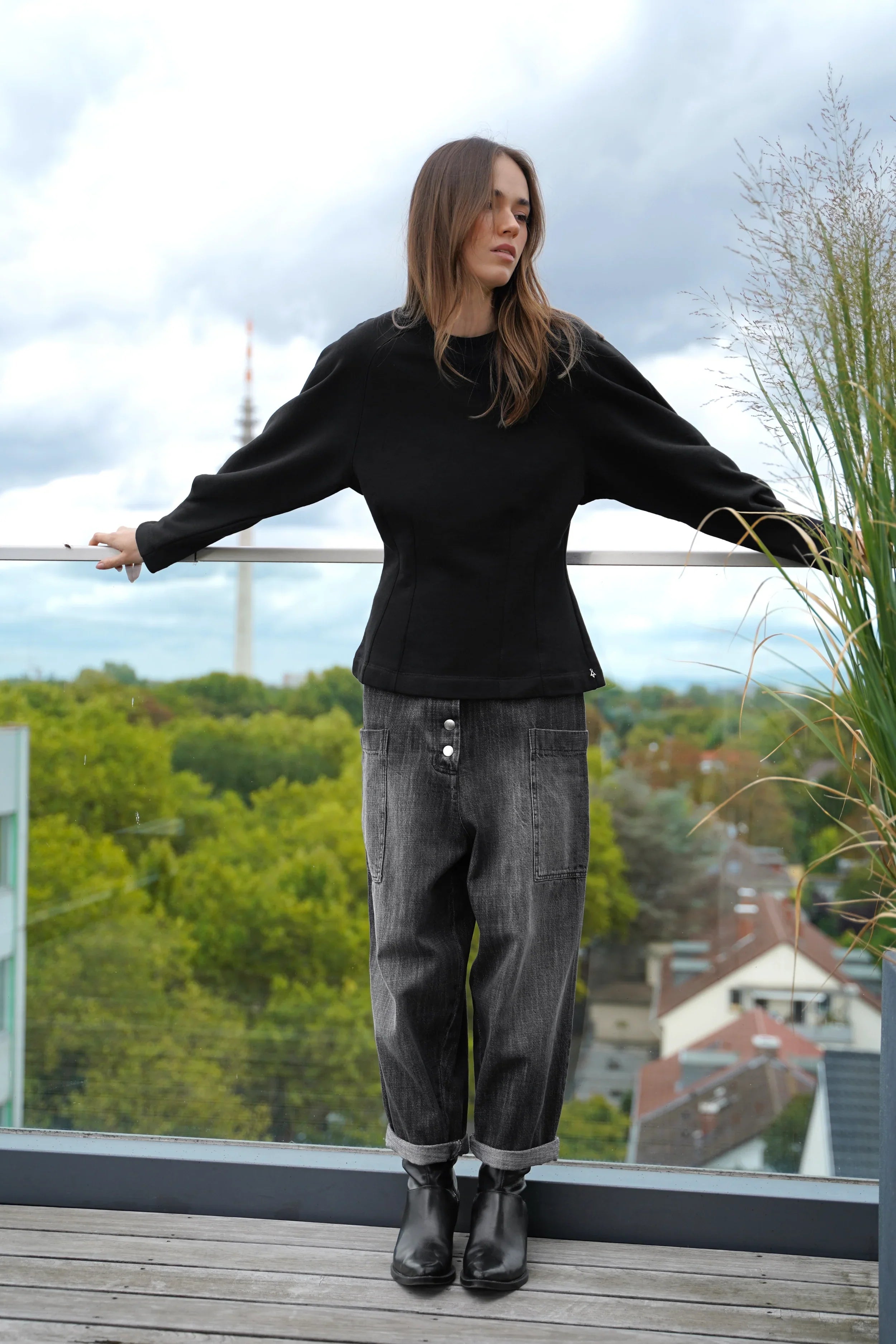 Woman wearing black MALVEA Sweater with relaxed fit and structured seams standing on balcony with city background