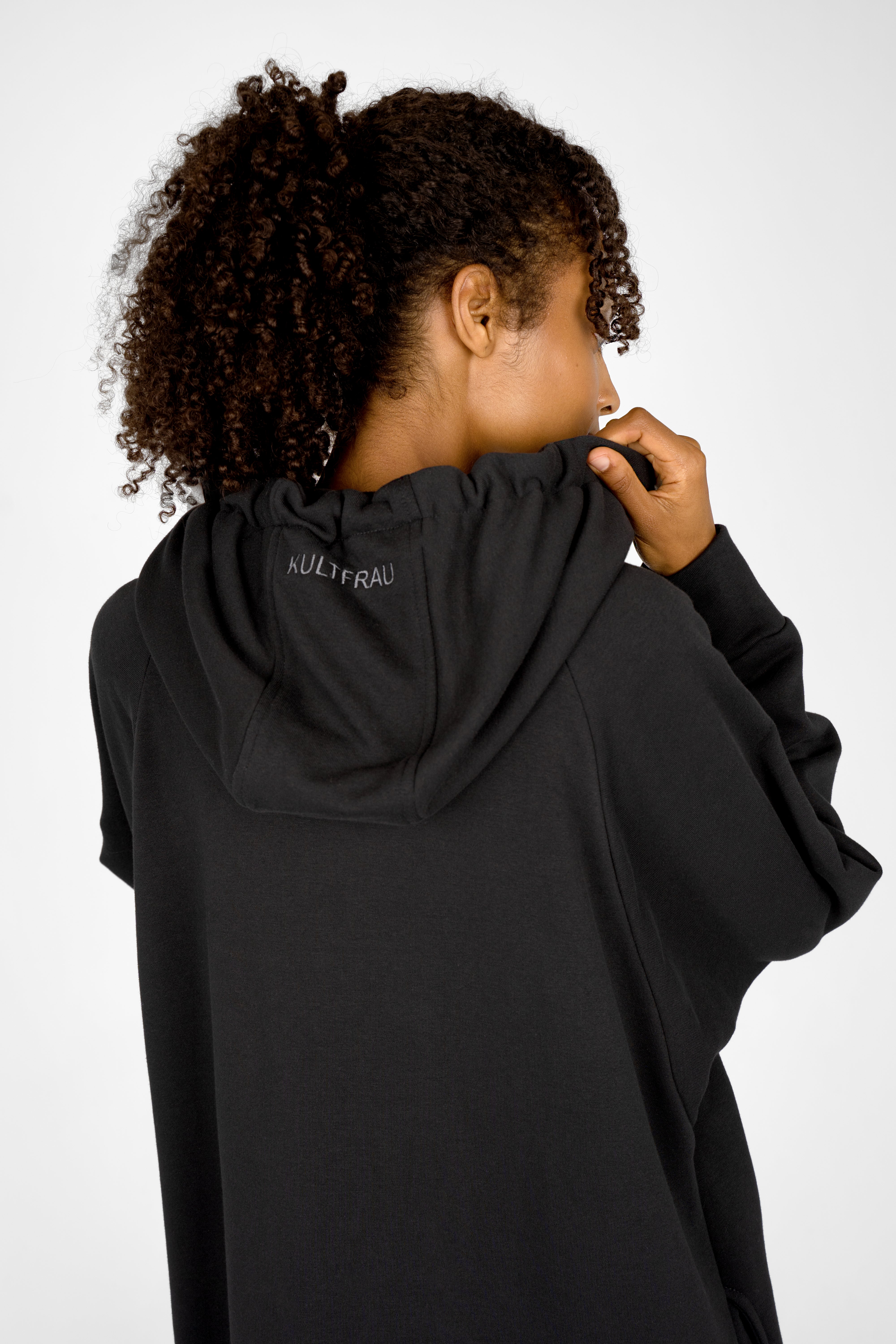 Back view of a woman wearing a black MANDI.K Longzip-Hoodie with a large hood and subtle logo embroidery, showing soft cotton fabric and relaxed fit.