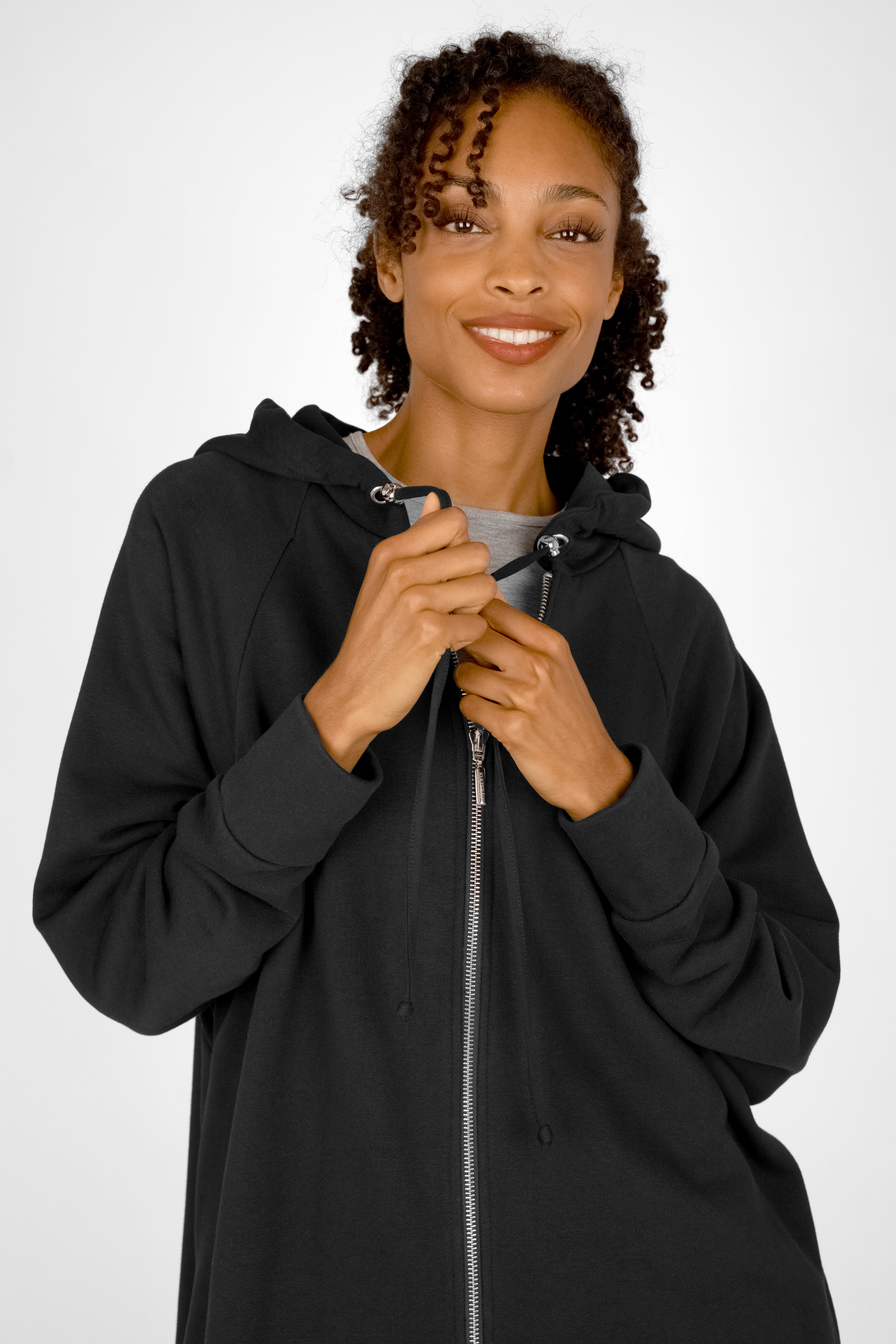 Woman wearing a black MANDI.K Longzip-Hoodie with a two-way zipper and hood, smiling and adjusting the zipper.