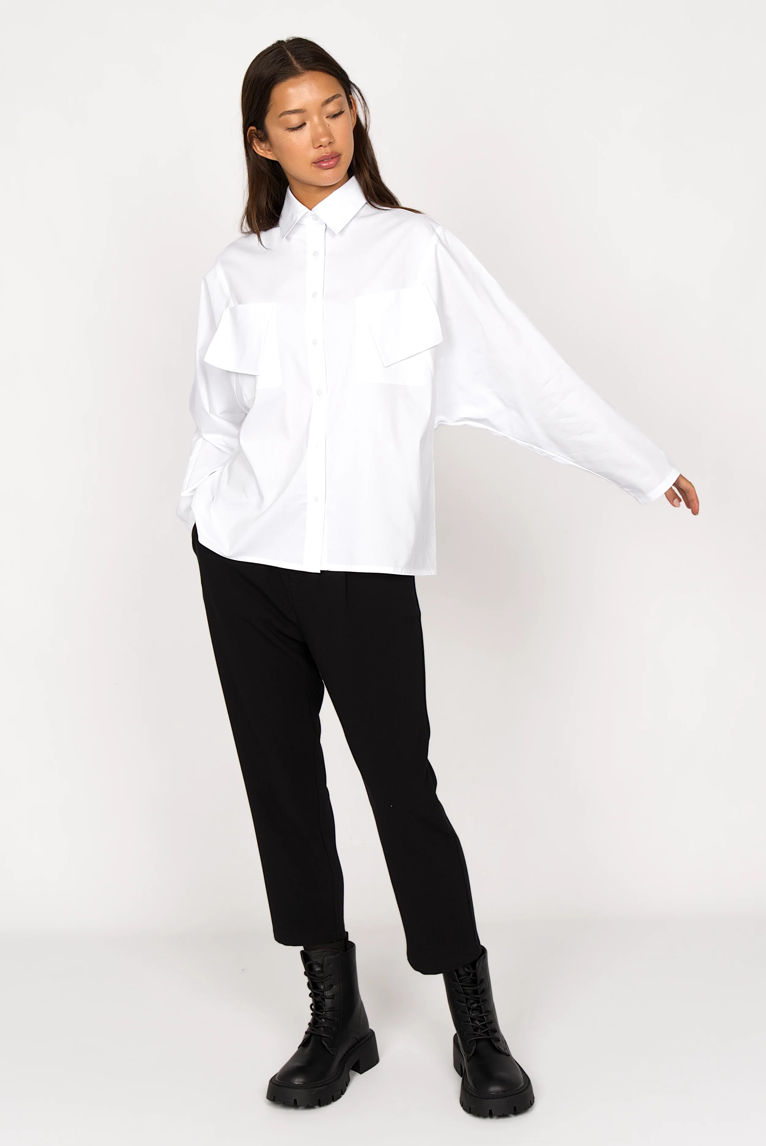 Woman wearing a white MELIZIA Bluse aus Popeline with bat-cut sleeves, asymmetrical pockets, and relaxed fit paired with black pants and boots