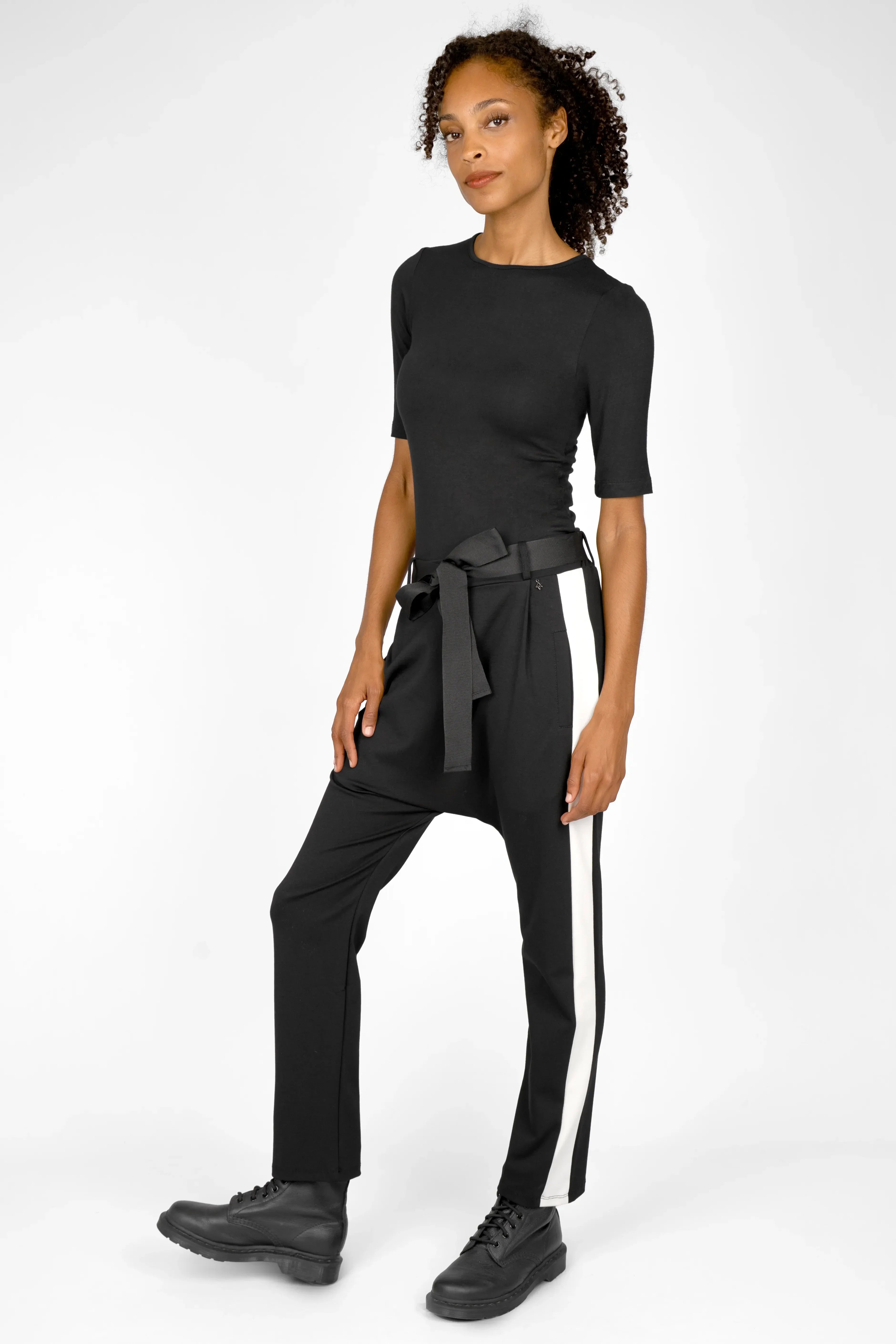 Frau trägt schwarze MYRAN Tapered Sweathose mit weißem Kontraststreifen, Bindegürtel und hochgekrempelten Beinen