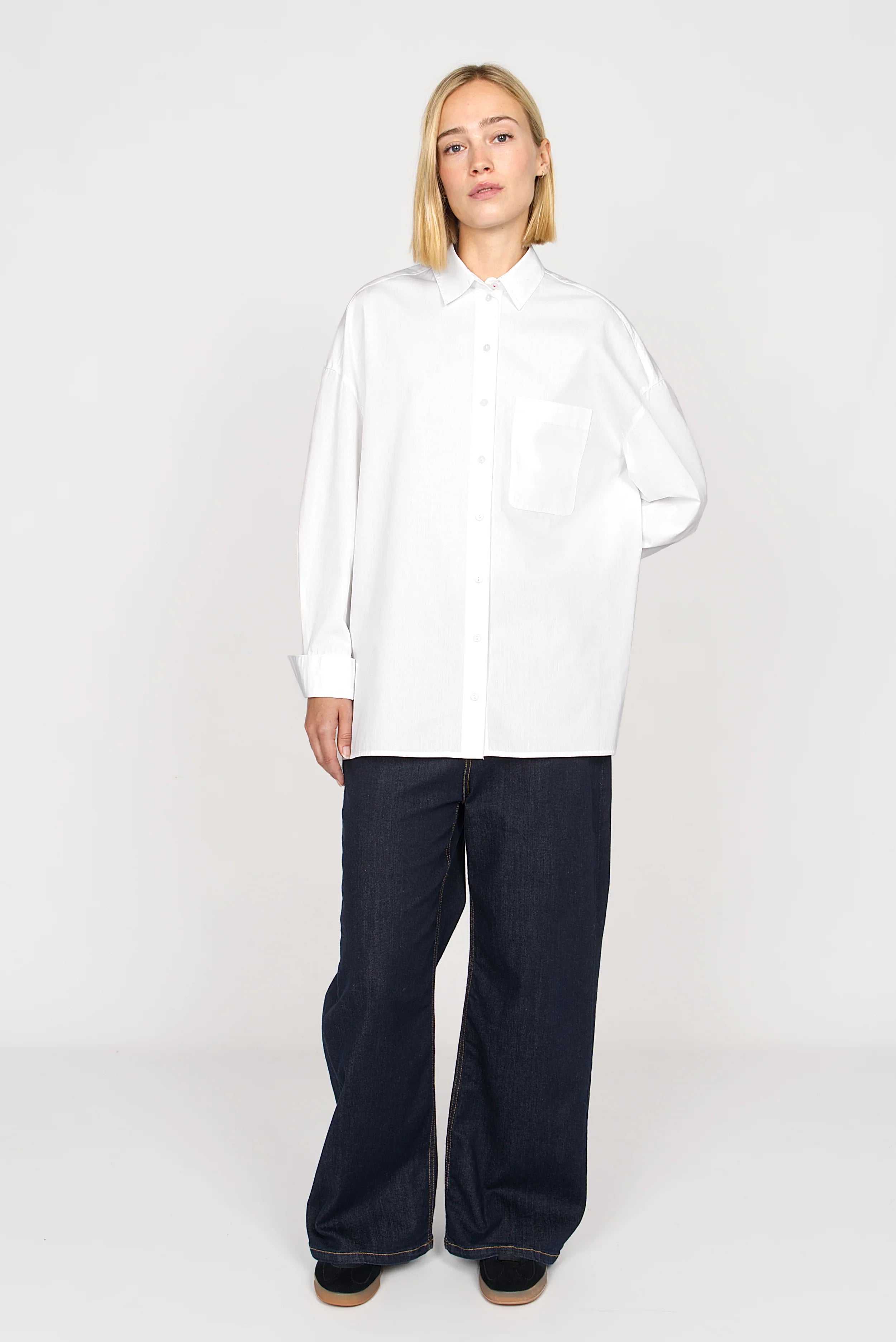 Woman wearing white NATI Statement Oversize poplin shirt with breast pocket and relaxed fit, paired with wide dark pants.