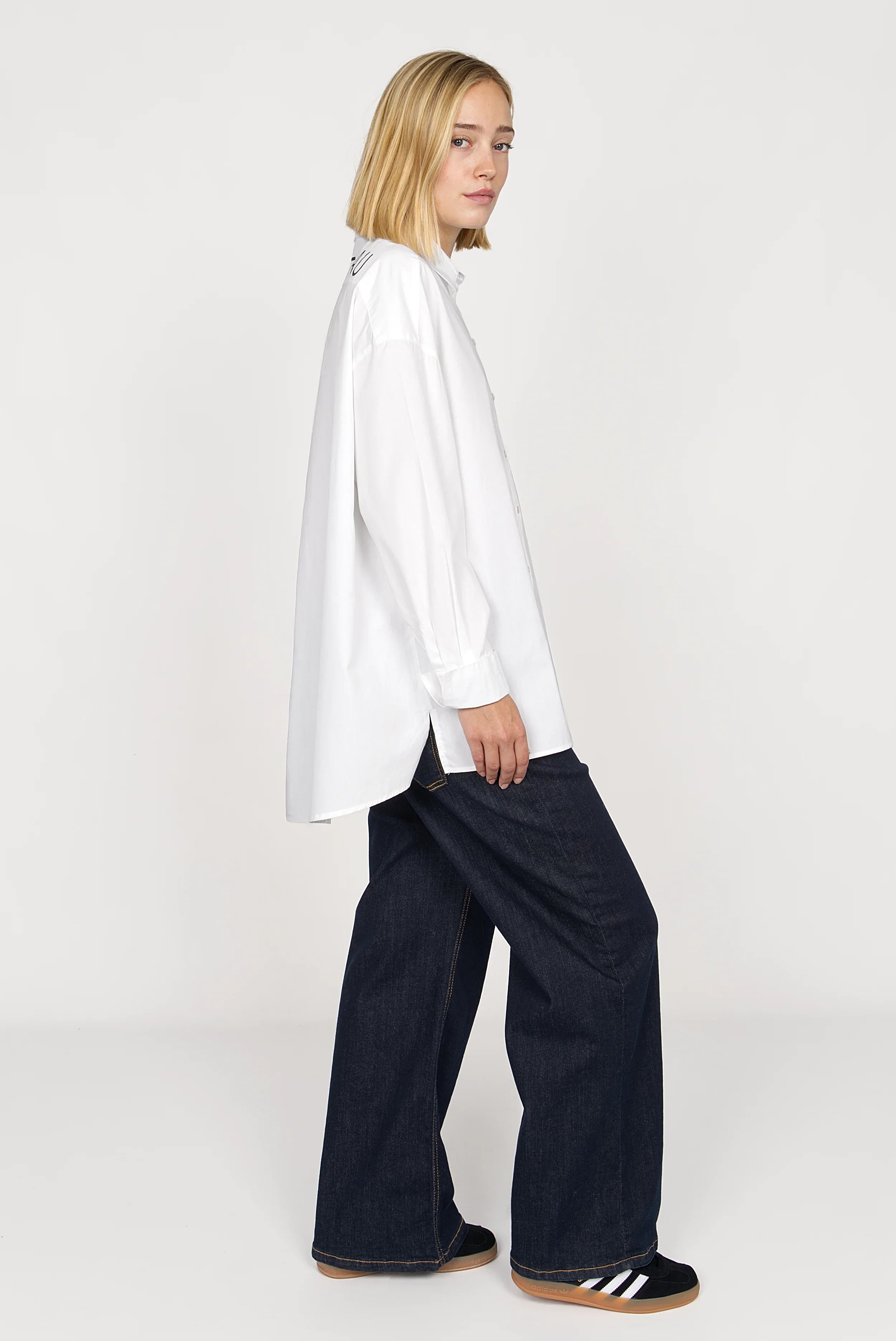 Woman wearing NATI Statement Oversize white popelin shirt with "KULTFRAU" print on back and wide cuffs, paired with dark wide-leg jeans and sneakers.