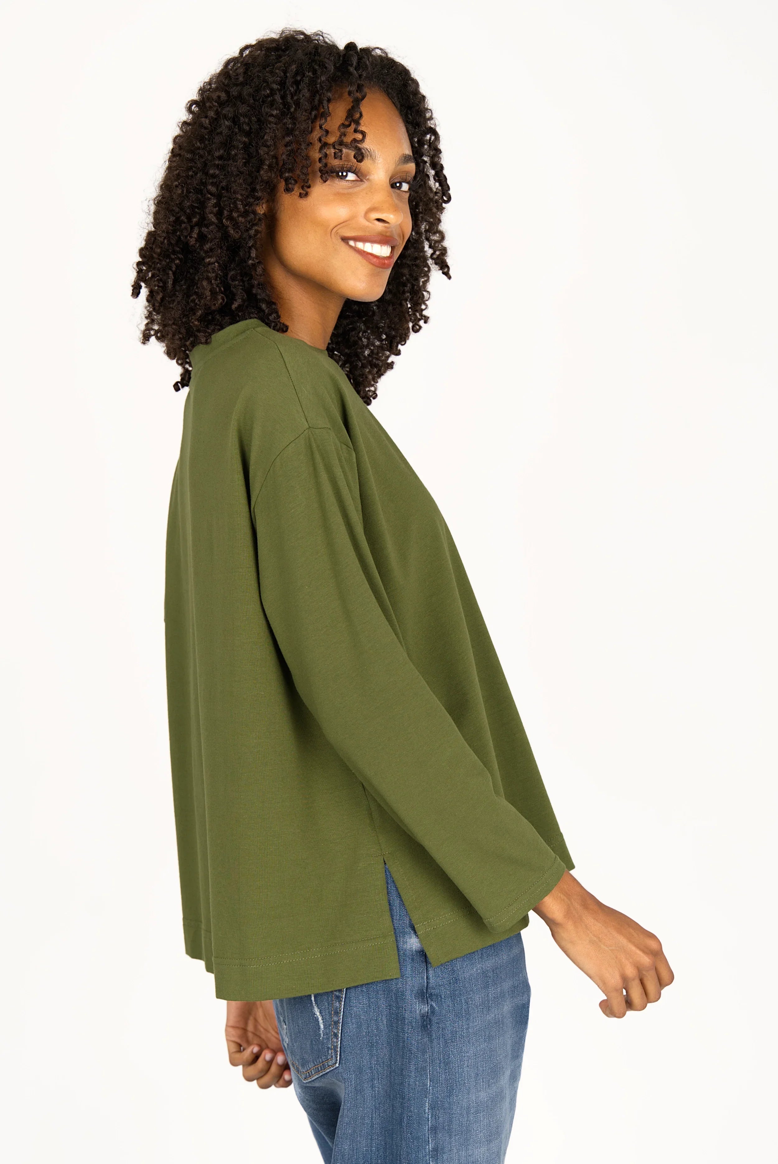 Woman wearing olive green NITI.K Langarm Shirt with relaxed fit and side slits, paired with blue jeans, smiling sideways view.