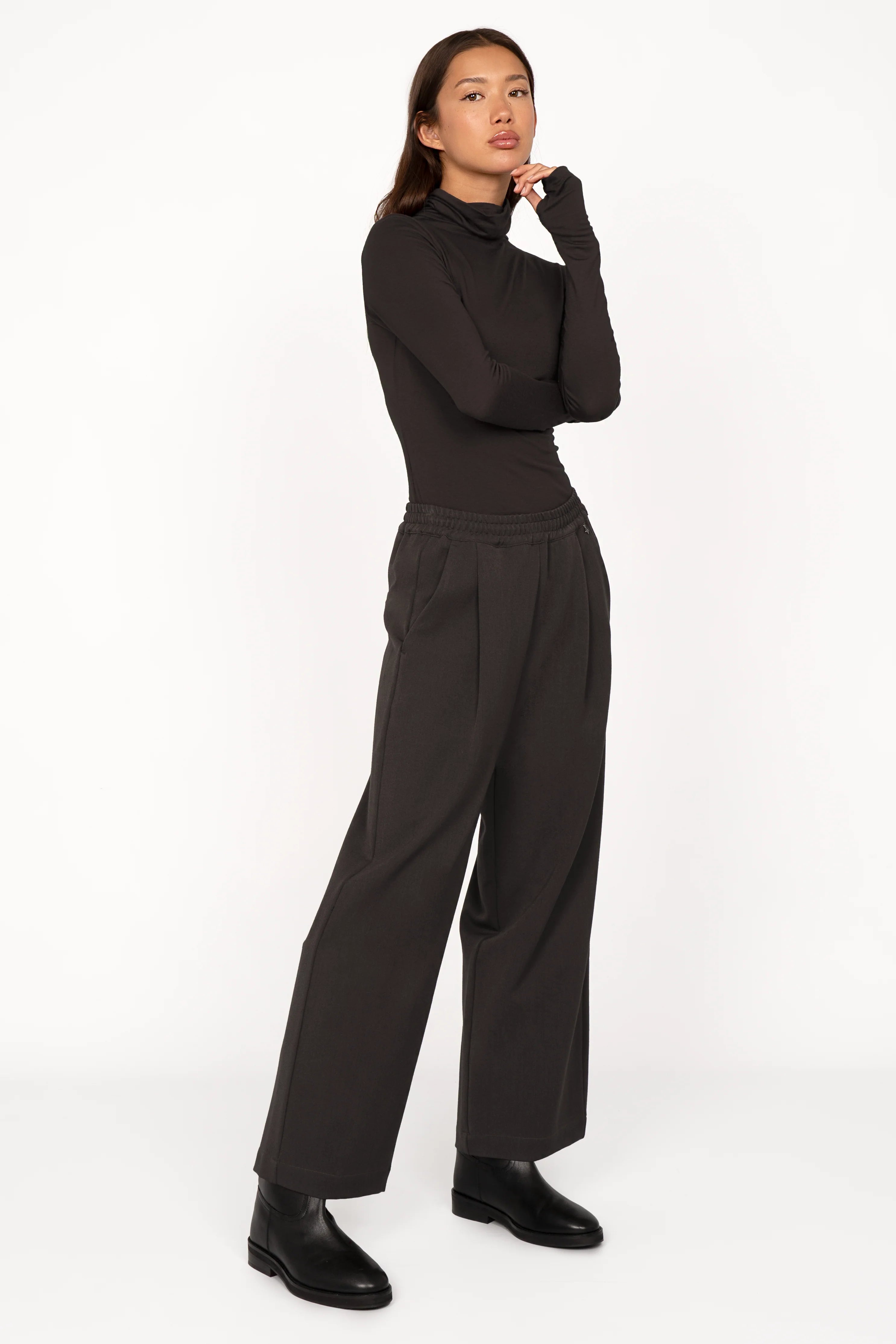 Woman wearing black ONA wide-leg hose with elastic waistband, side pockets, and relaxed fit, styled with black turtleneck and boots against white background