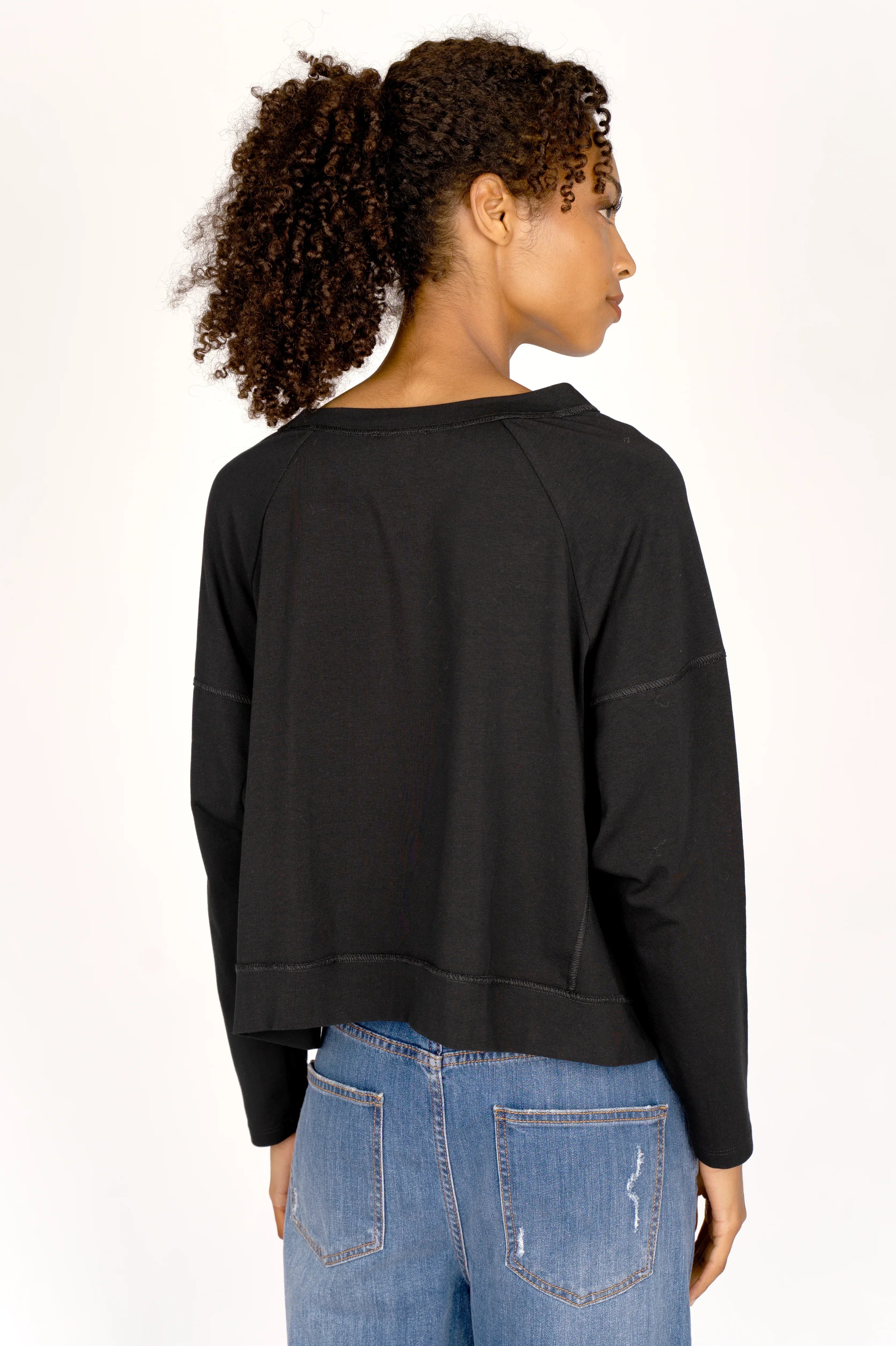 OSASU Cropped Longsleeve schwarzes, locker geschnittenes Langarmshirt mit weitem Rundhalsausschnitt und geschnittenen Kanten, getragen von einer Frau von hinten sichtbar