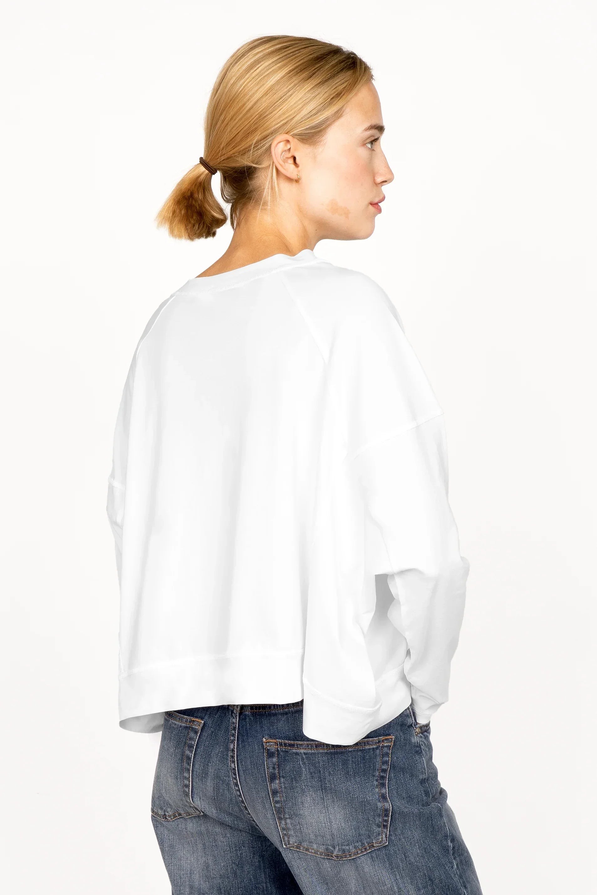 Frau trägt OSASU Cropped Longsleeve mit weitem Rundhalsausschnitt und locker fallender Kastenform, weißes Shirt hinten länger geschnitten.