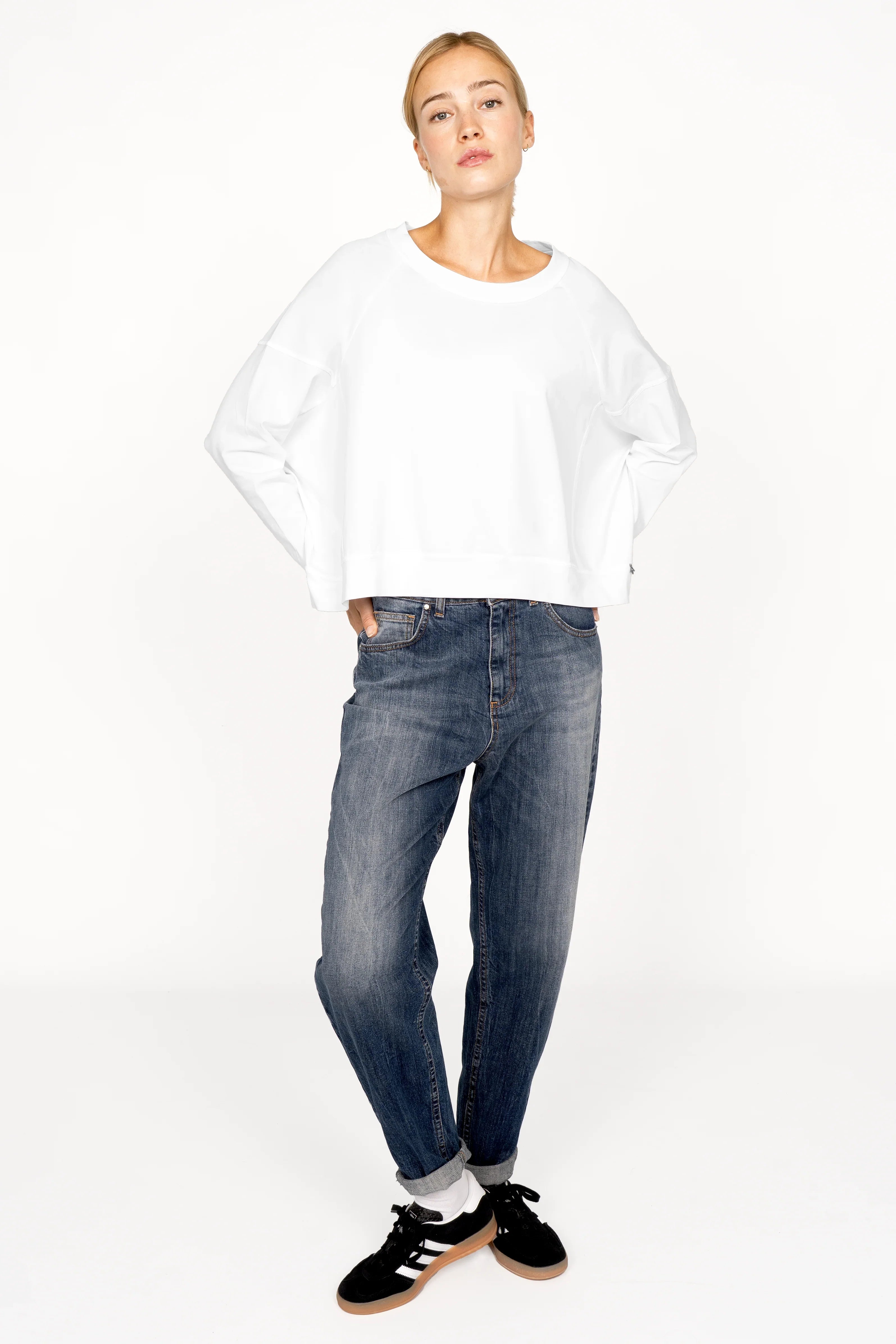 Frau trägt weißes OSASU Cropped Longsleeve mit weitem Rundhalsausschnitt und Kastenform, kombiniert mit Jeans und Sneakern.