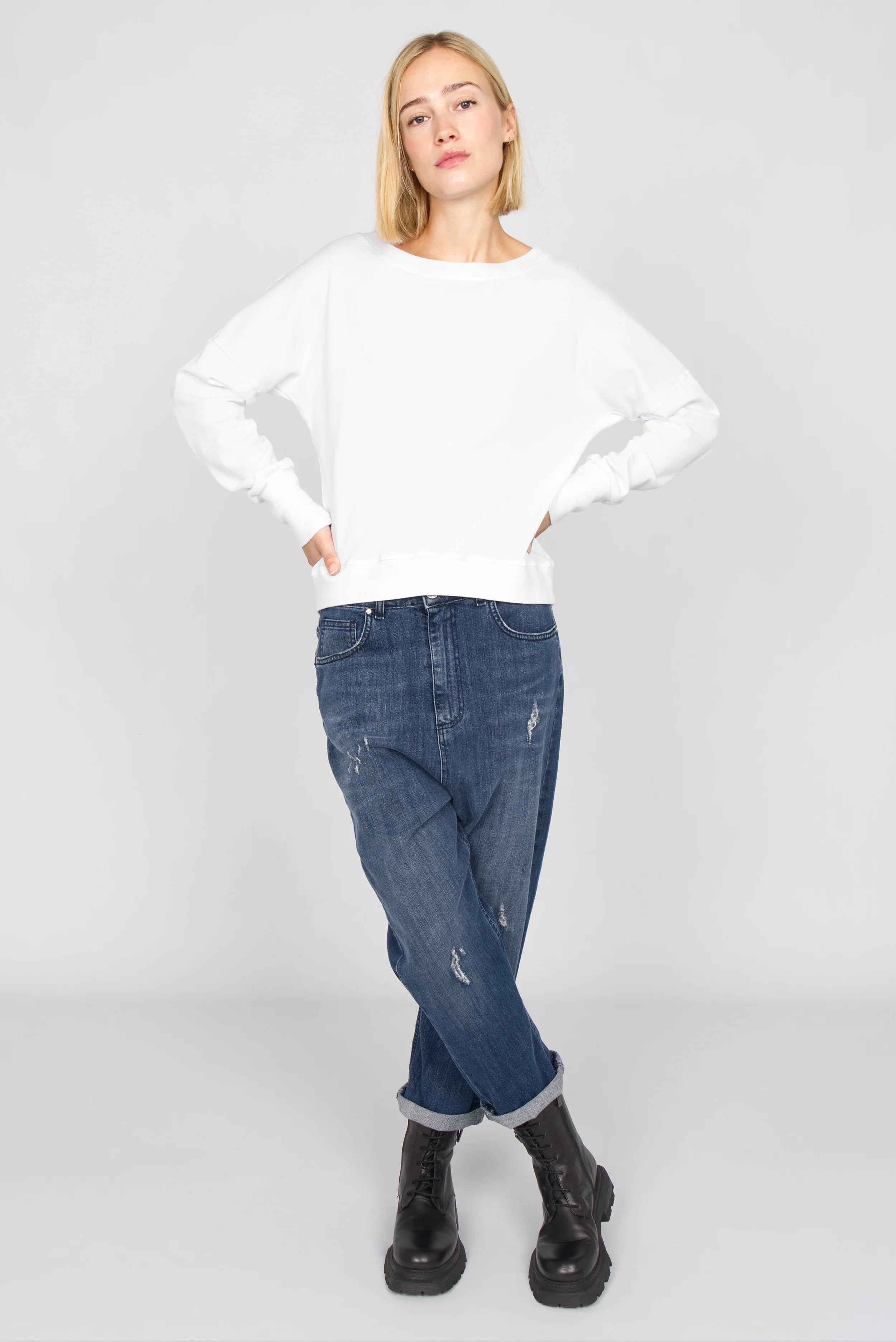 Frau trägt weißes RIISA Longsleeve mit U-Boot-Ausschnitt, überschnittenen Schultern und extra breiten Rippbündchen zu blauen Jeans und schwarzen Stiefeln.