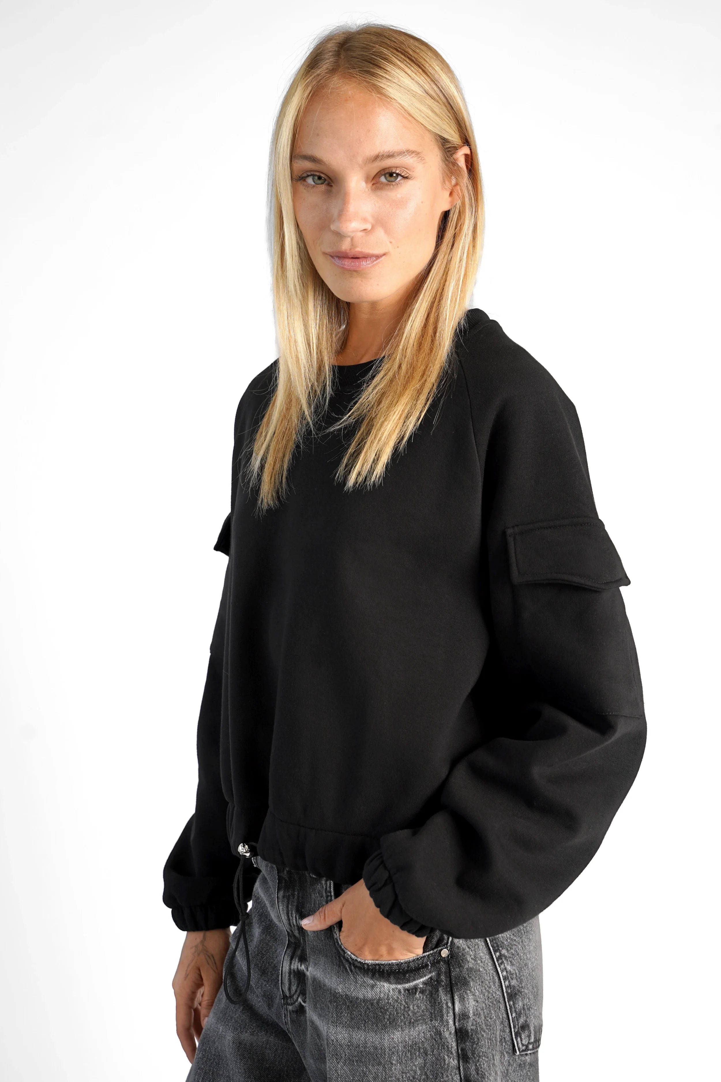 Woman wearing black SEATA cropped sweater with oversized fit, round neckline, long sleeves with elastic cuffs, front flap pockets, and drawstring hem.