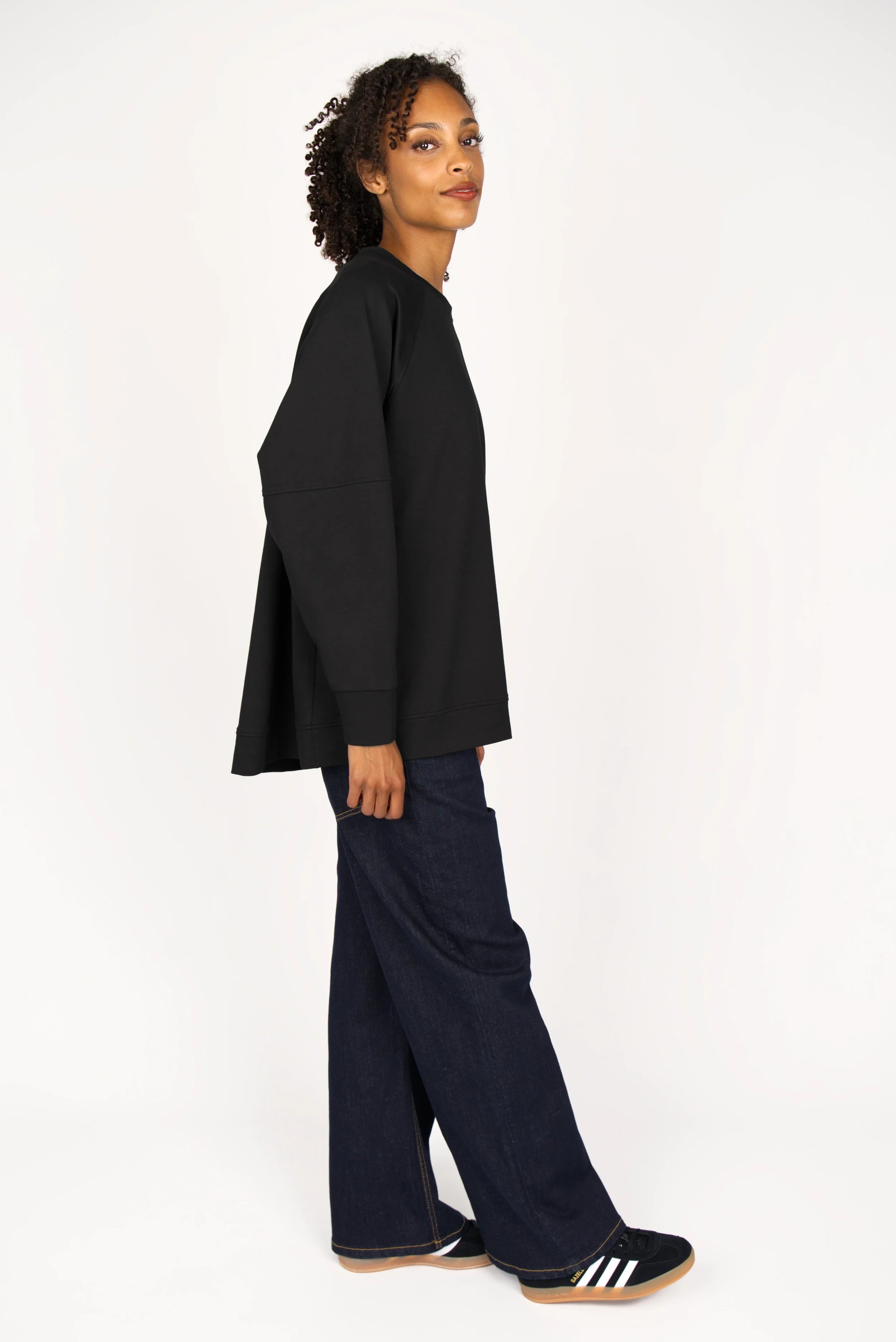 Black SELKA Sweater with A-line silhouette, raglan sleeves, and longer back hem on model wearing dark pants and sneakers