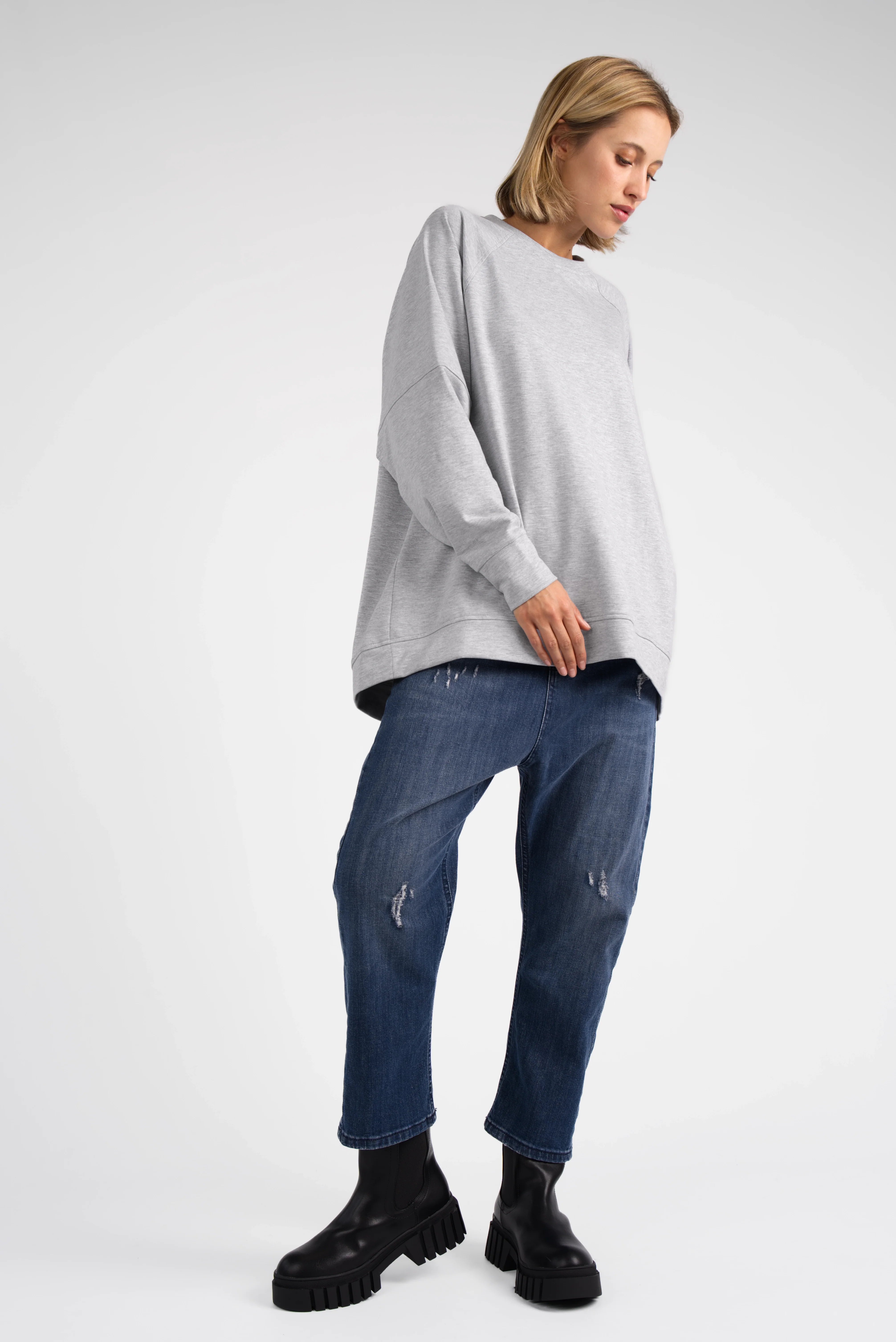 Woman wearing a grey SELKA Sweater with A-line shape, raglan sleeves, and longer back hem paired with blue distressed jeans and black boots.