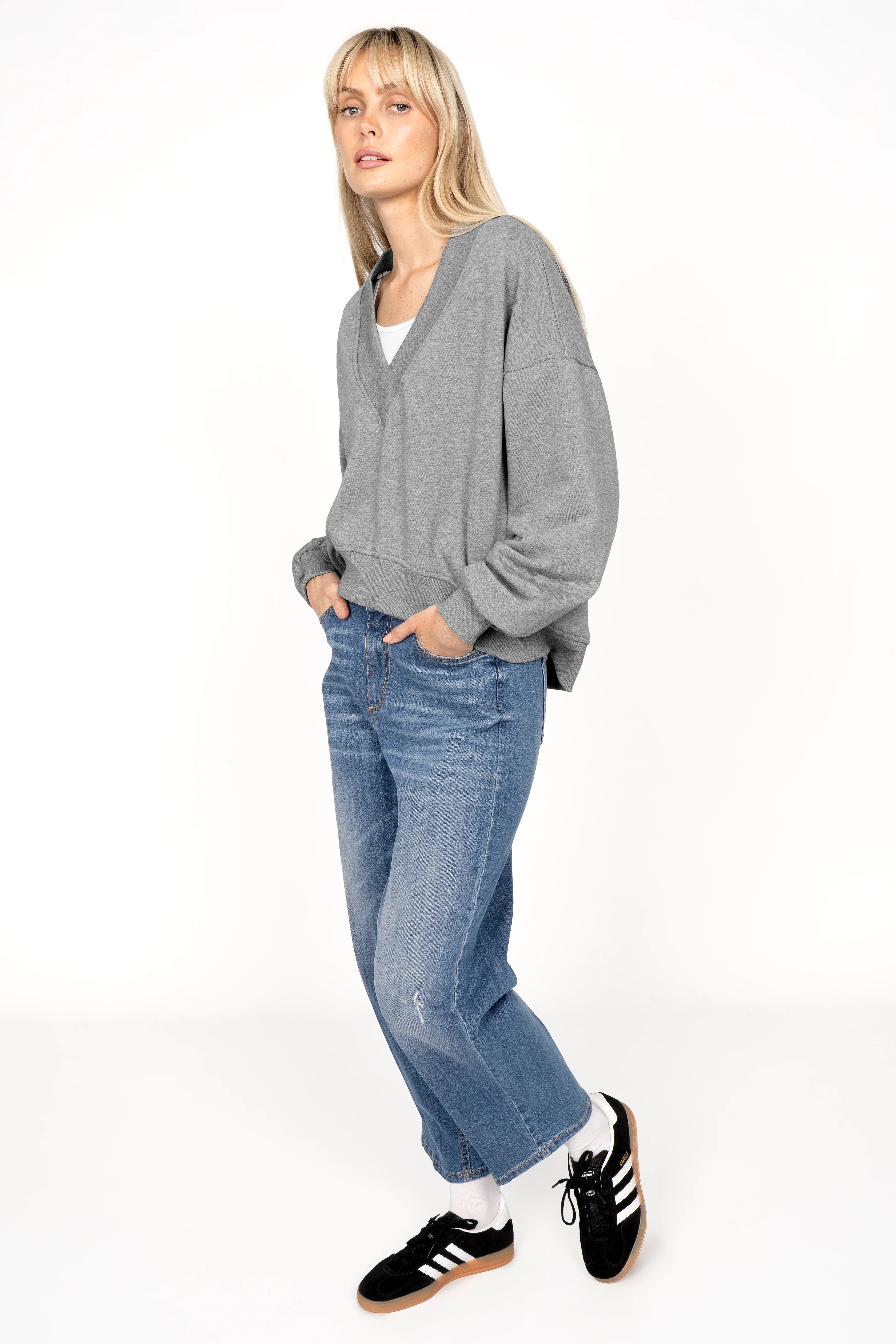 Woman wearing grey SEREA Sweater with deep V-neck, relaxed fit, and cuffed sleeves paired with blue jeans and black sneakers.
