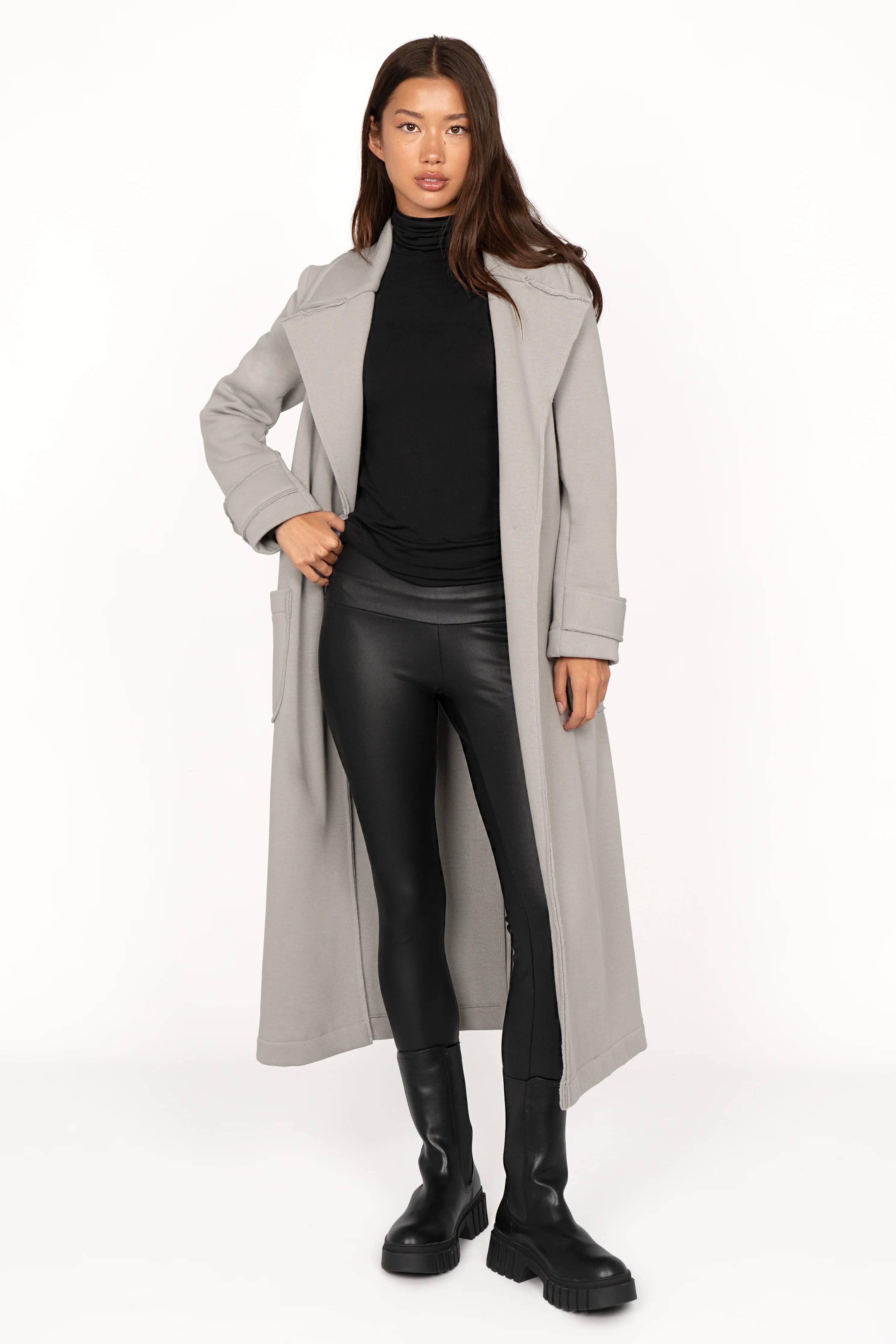 Woman wearing SIMPI Leggings made of stretchy eco-leather with a wide waistband, paired with a long grey coat and black boots.