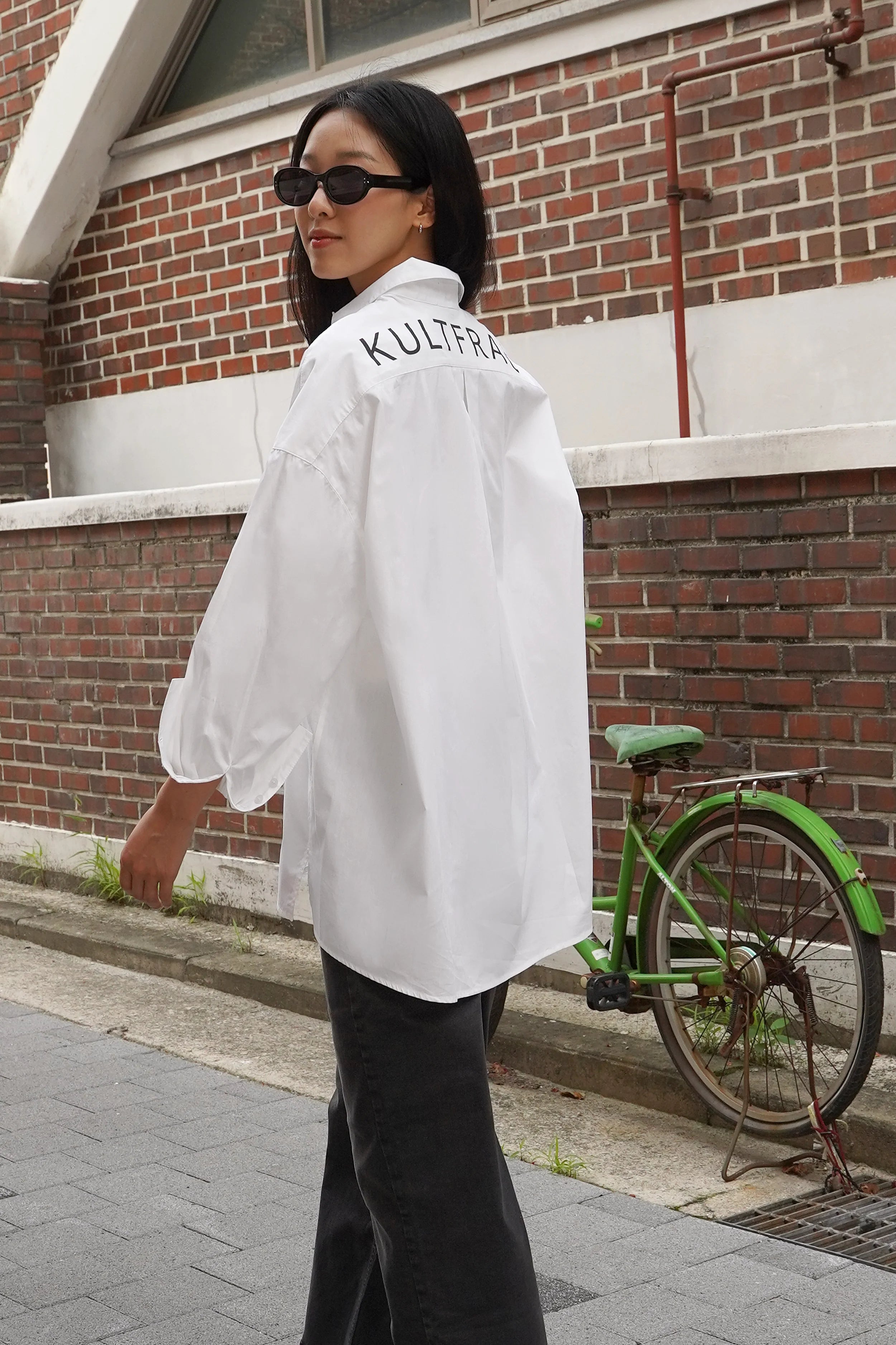 Woman wearing NATI Statement Oversize white popelin shirt with "KULTFRAU" print on back, standing outdoors near brick wall and green bicycle