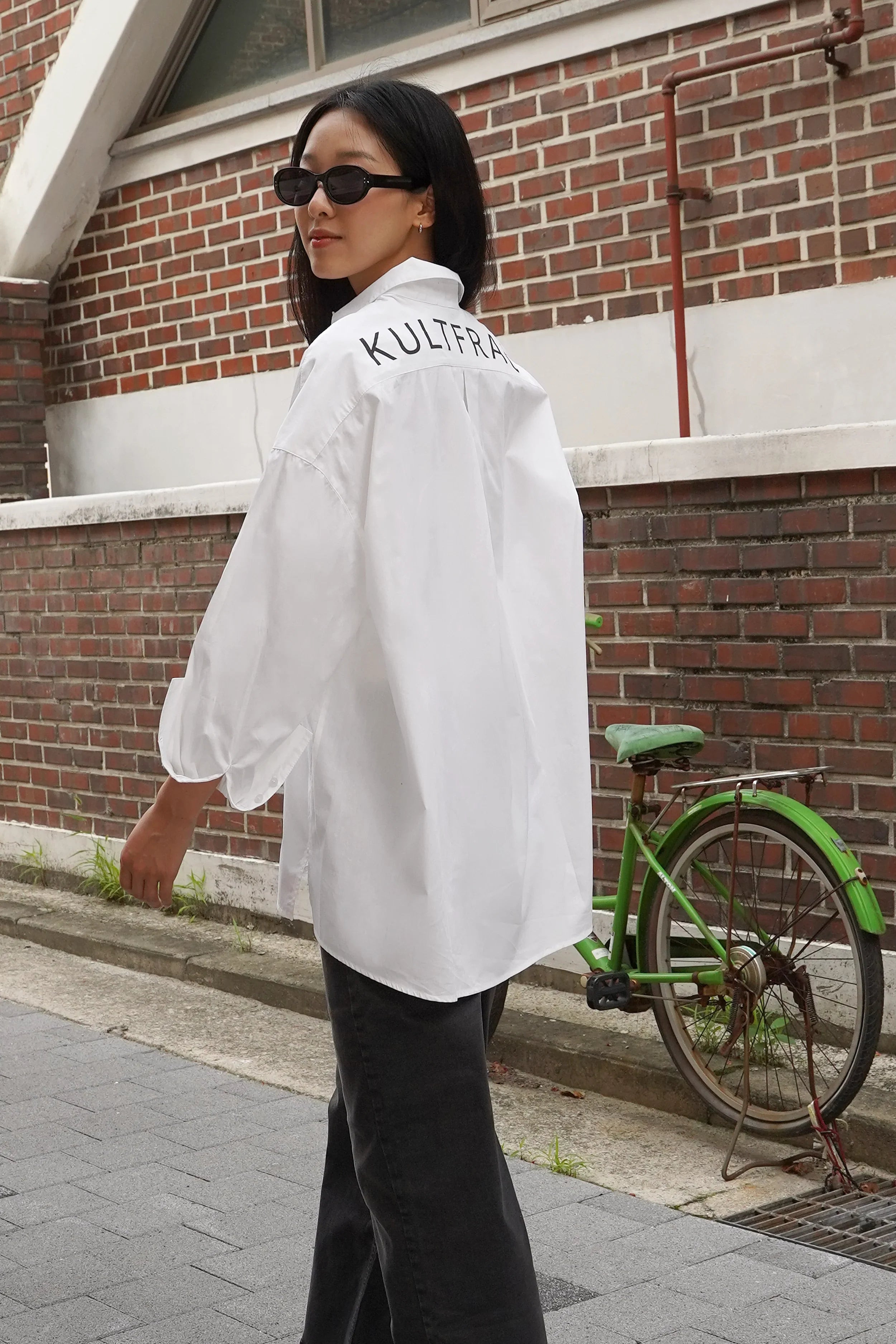 Woman wearing NATI Statement Oversize white popelin shirt with "KULTFRAU" print on back, standing outdoors near brick wall and green bicycle