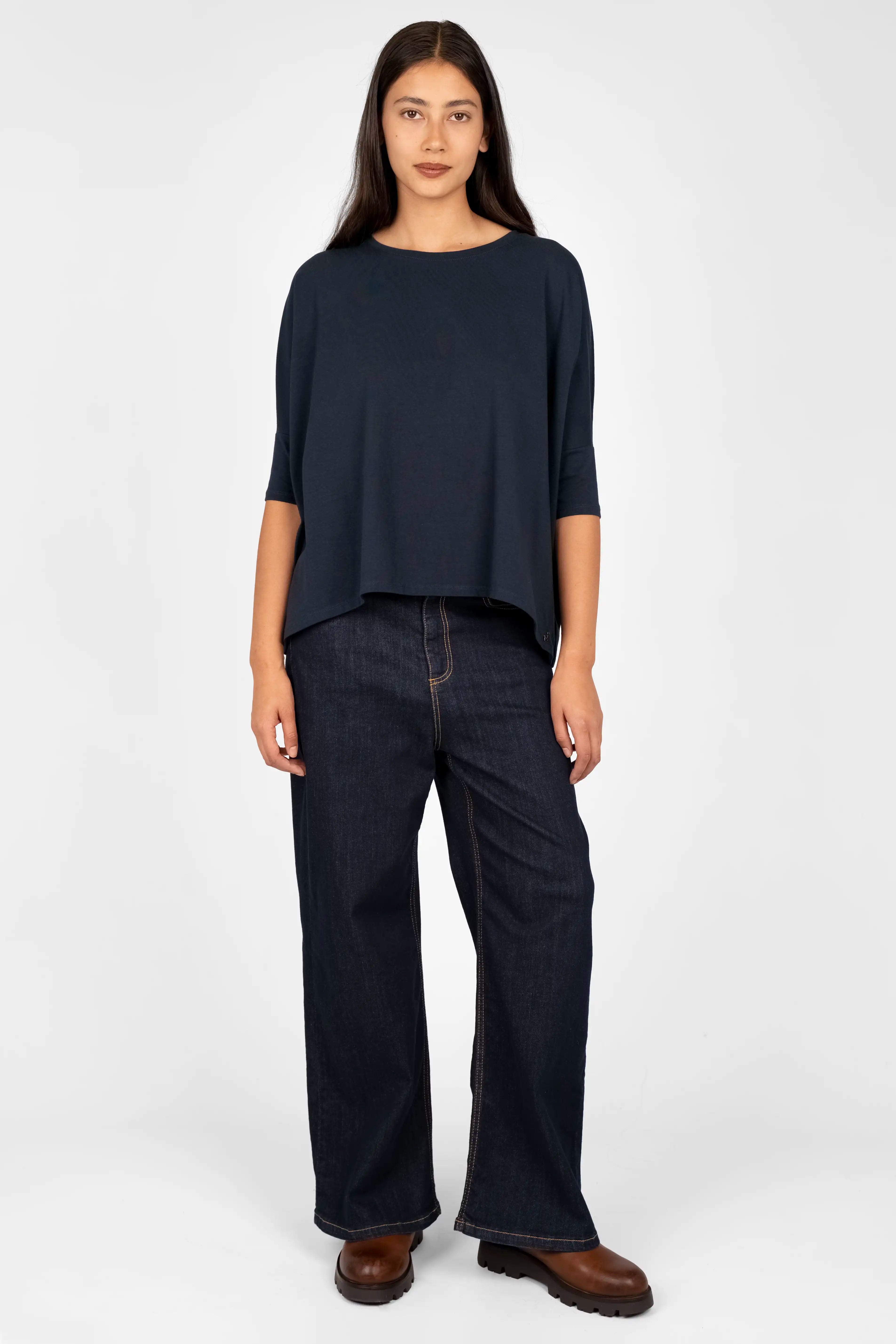 Damen trägt dunkelblaues TAMI.K Blusenshirt in weiter Kastenform mit Rundhals und leicht abgerundetem Saum, kombiniert mit dunkler Hose und braunen Schuhen.