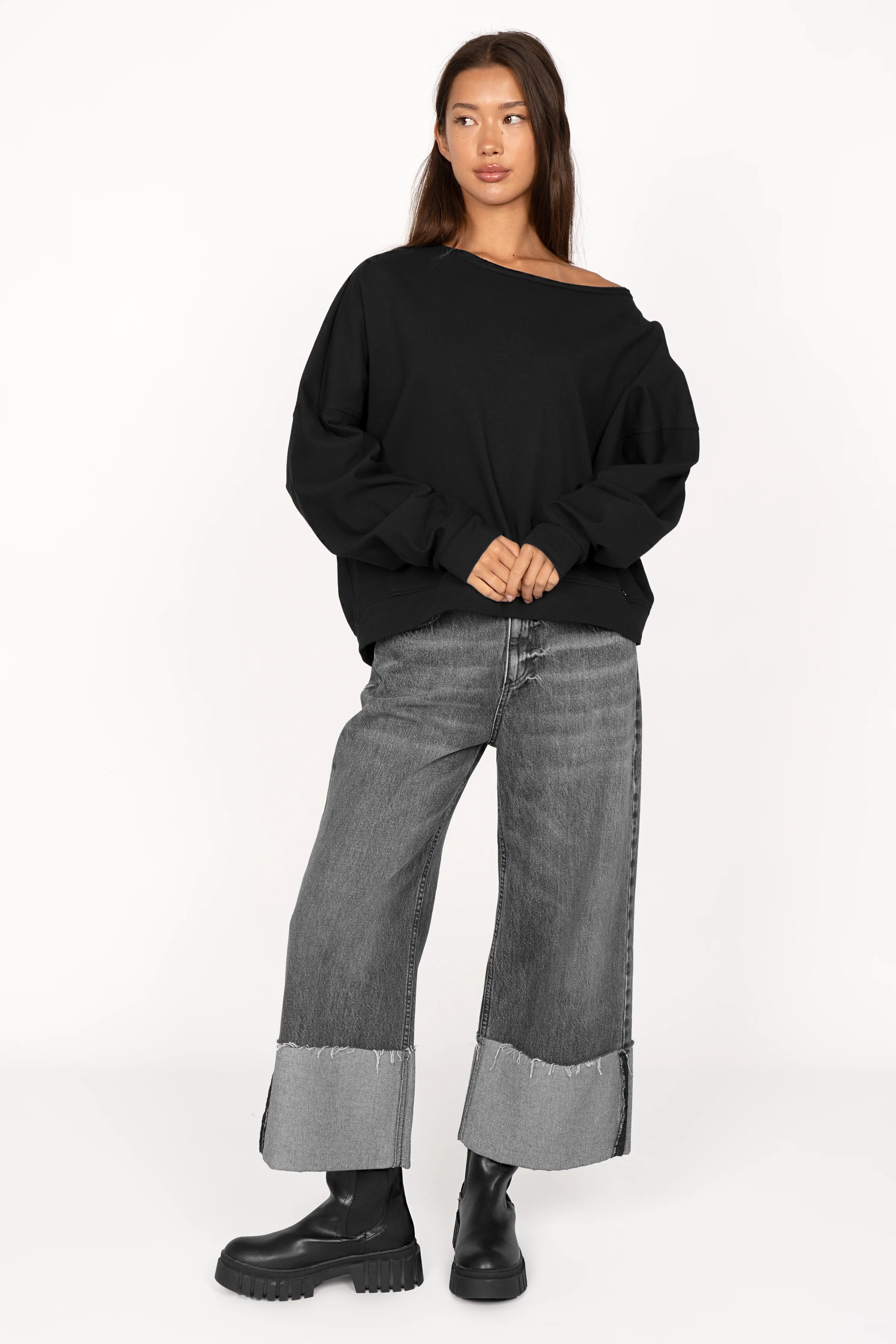 Woman wearing black TIMAJ one shoulder sweater with wide neckline, loose fit, and extended sleeves paired with wide cuffed gray jeans and black boots