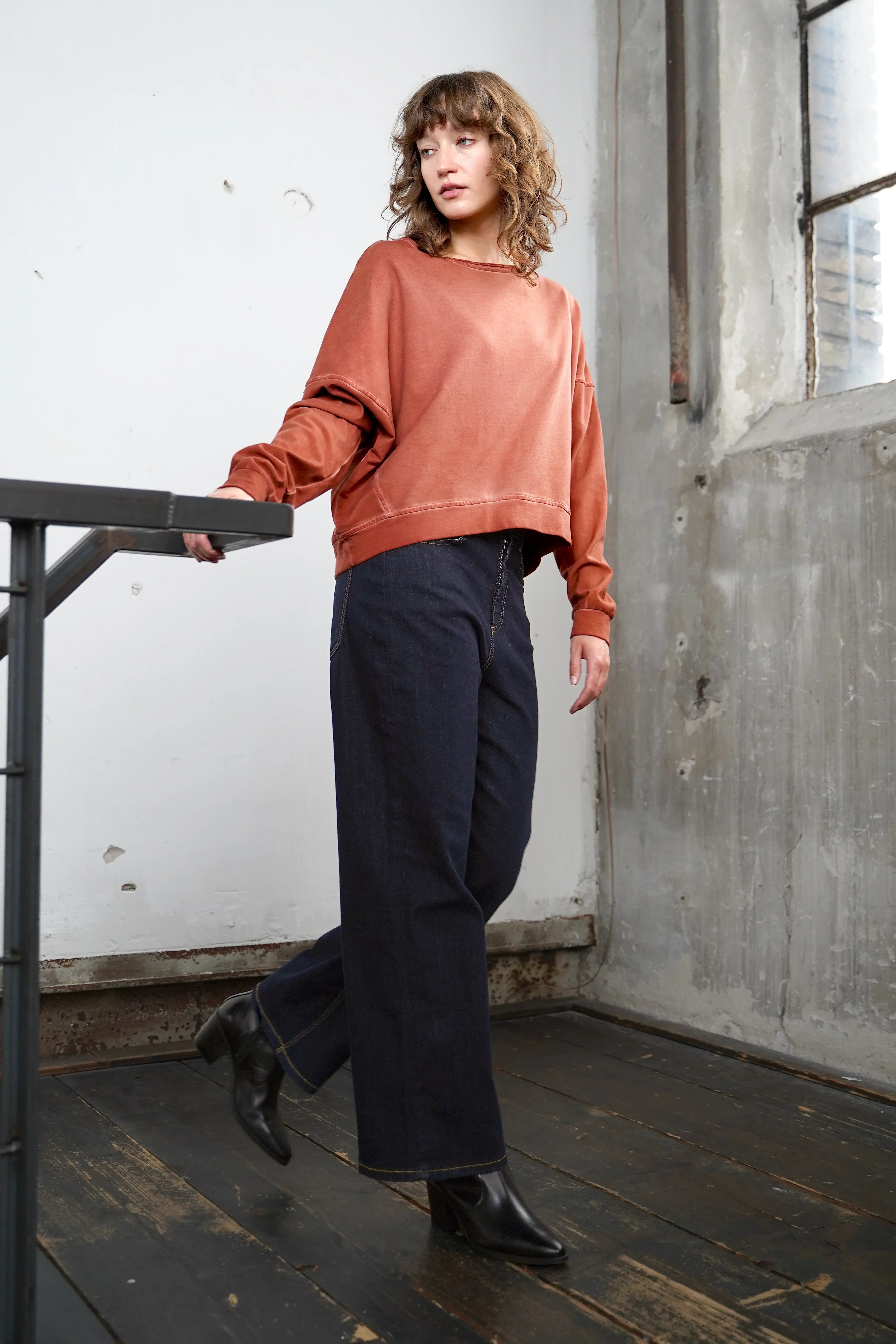 Women wearing TIMAJ Revive One Shoulder Sweater in mocha vintage wash with wide neckline and loose fit paired with black pants in industrial setting