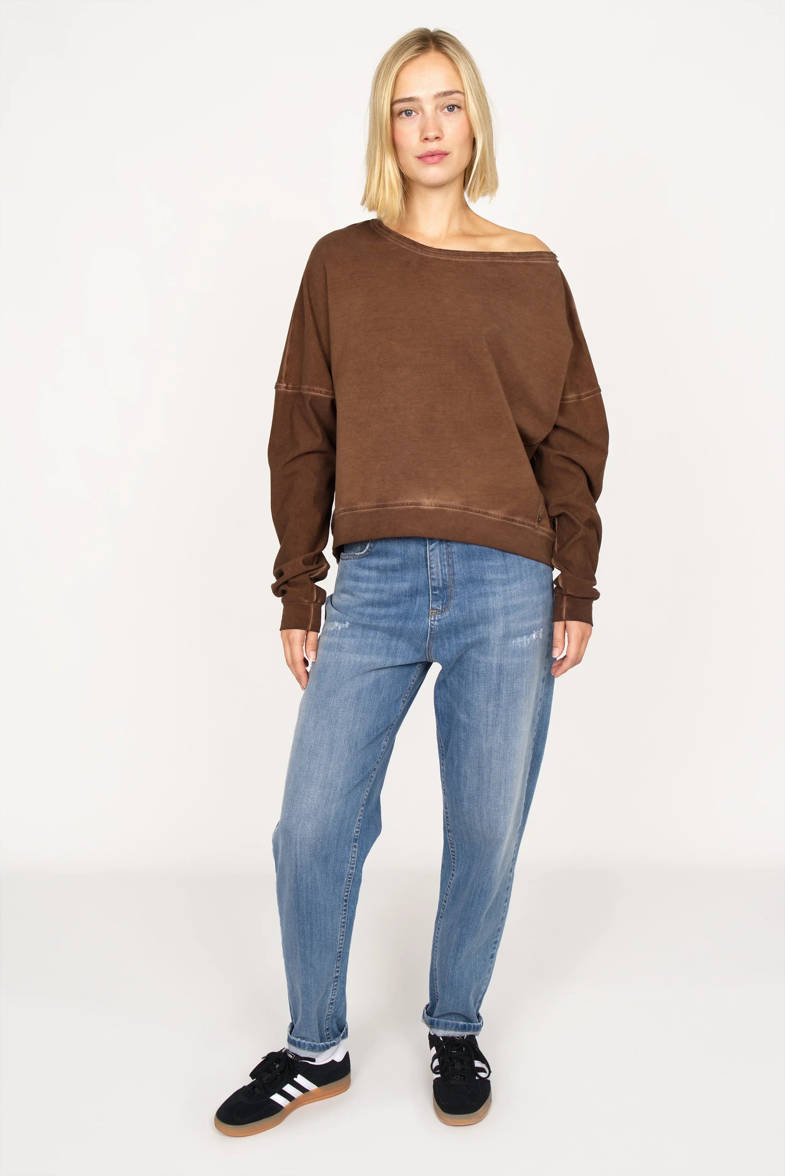 Woman wearing a mocha TIMAJ Revive One Shoulder Sweater with vintage wash and loose fit paired with blue jeans and black sneakers.
