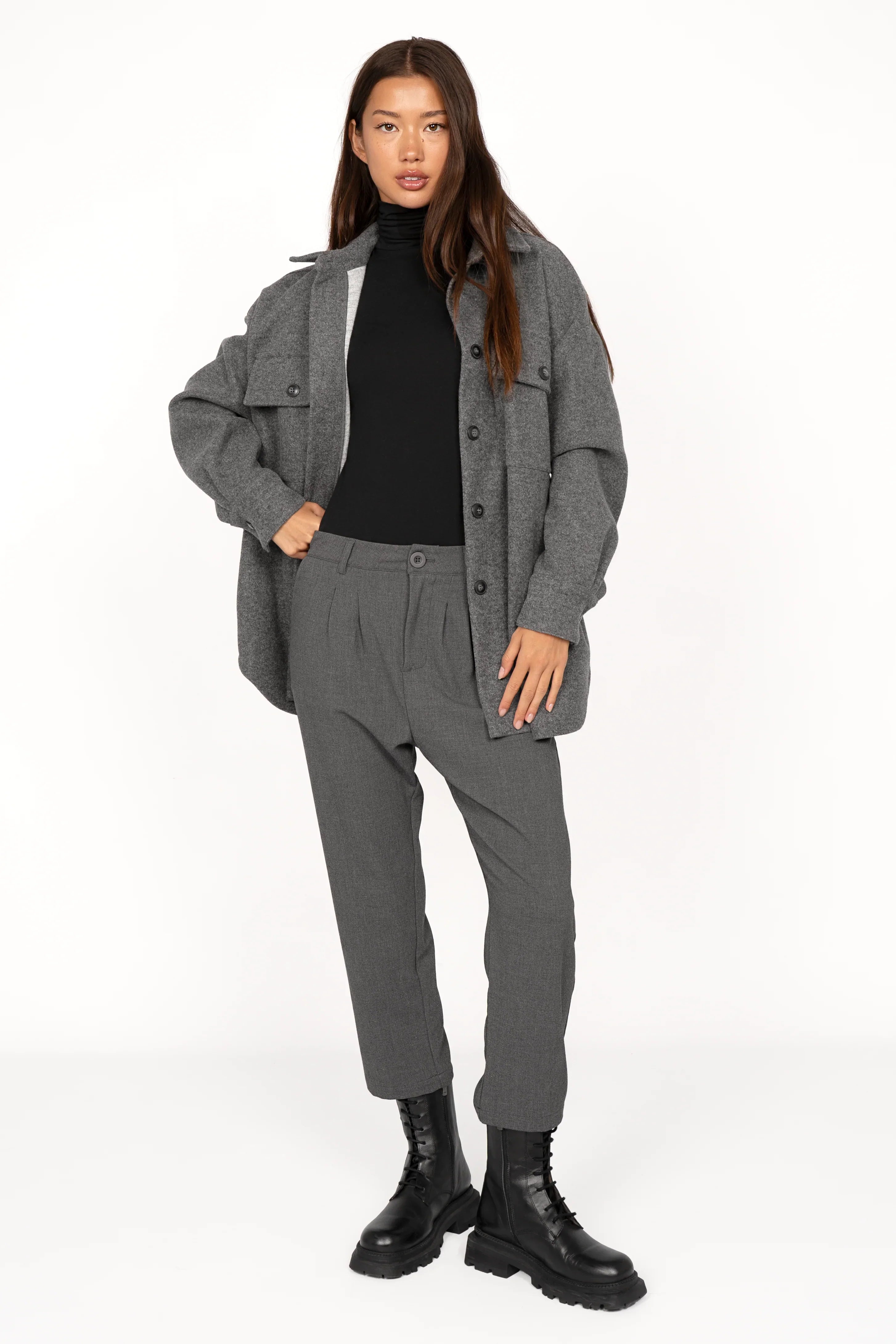 Frau trägt graue TINKE Hose Cropped Fit mit Bundfalten, tiefem Schritt und Reißverschluss, kombiniert mit schwarzem Rollkragenpullover und grauer Jacke