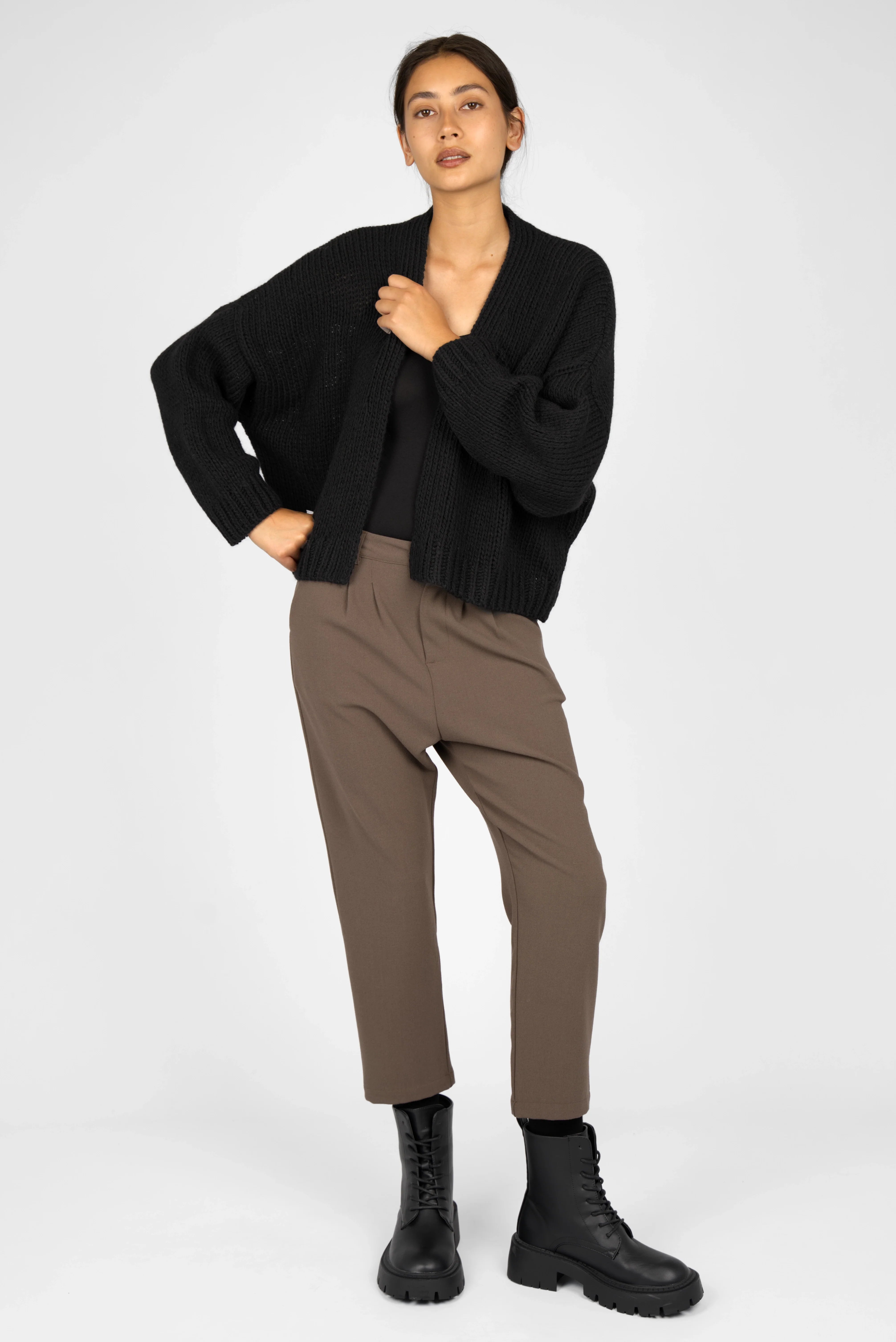 Woman wearing TINKE Hose Cropped fit brown trousers with cropped length, light pleats, and a relaxed fit paired with black boots and a black cardigan